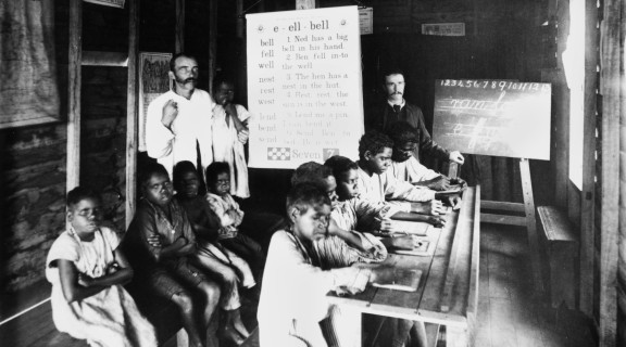 Side view of two rows of First Nations children sitting in a classroom with two men standing at the back at the end of the rows.