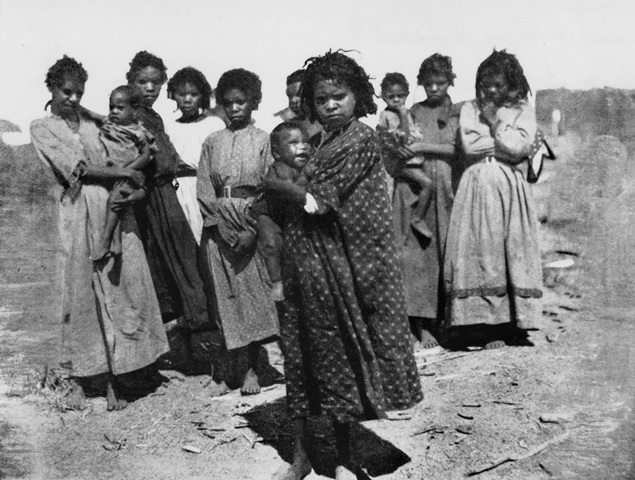 Photo from 1916 depicting a group of Indigenous women and children standing in long old fashioned dresses, squinting into the camera in bright sunlight.