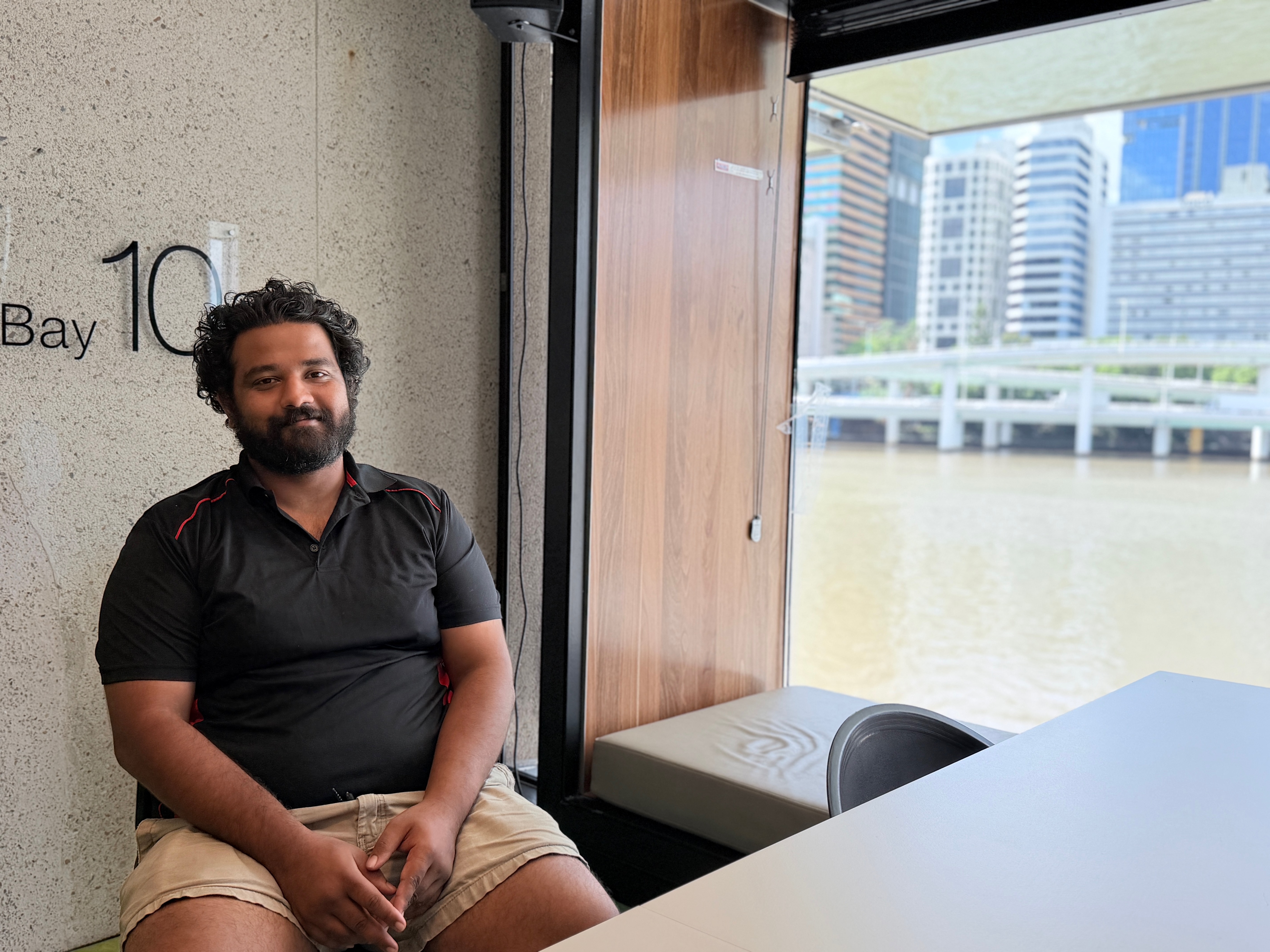 Amal sits on a chair with his arms in his lap, he is wearing a dark grey polo shirt and tan shorts, he is seated in front of a window that overlooks a section of the Brisbane river with the some skyscapers in the background. He has dark curly hair and a beard, he is smiling slightly at the camera.