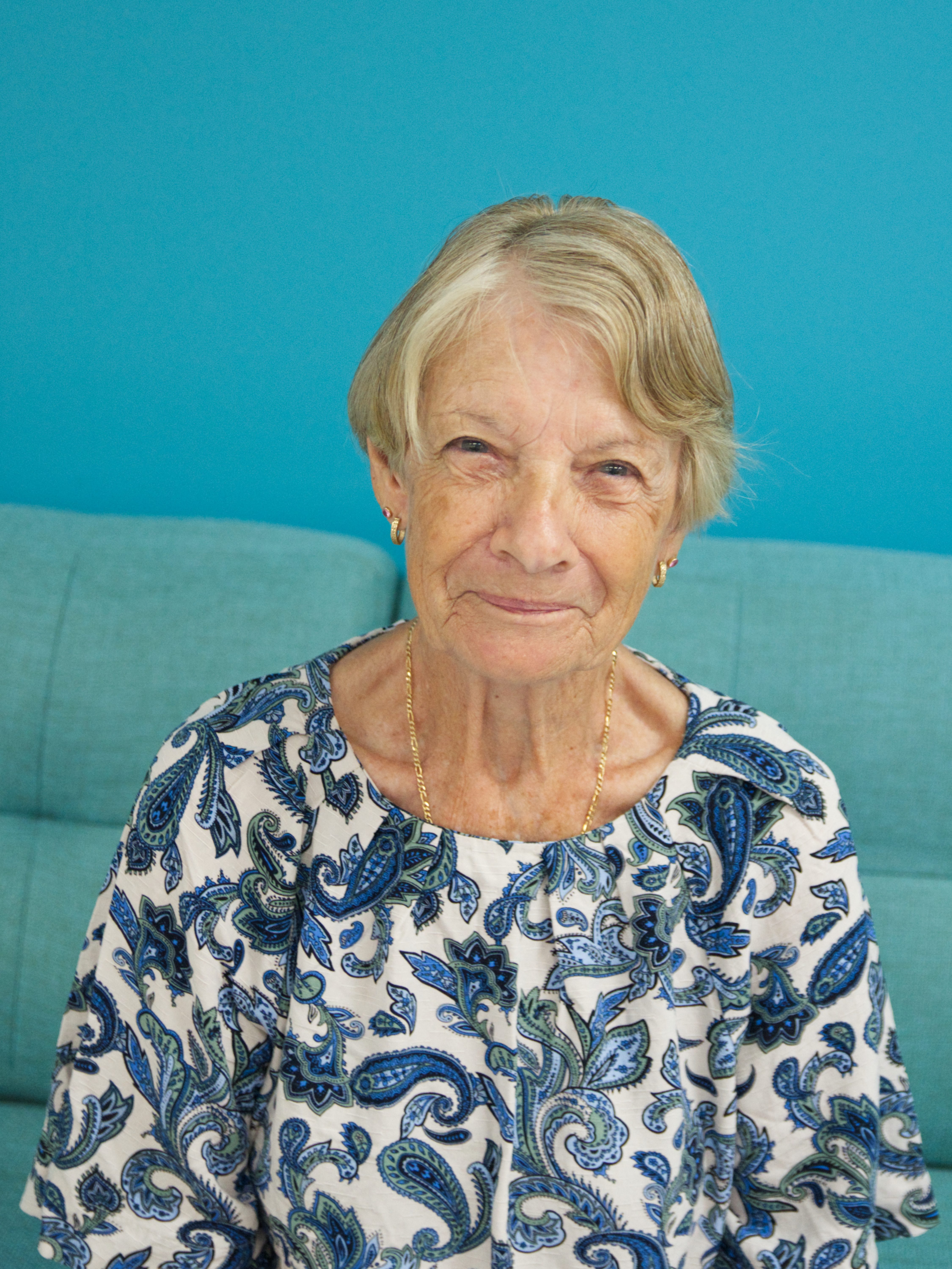 portrait photograph of a woman with short grey hair, gold earrings and necklace, a paisley blue pattern shirt, smiling at the camera with a blue background