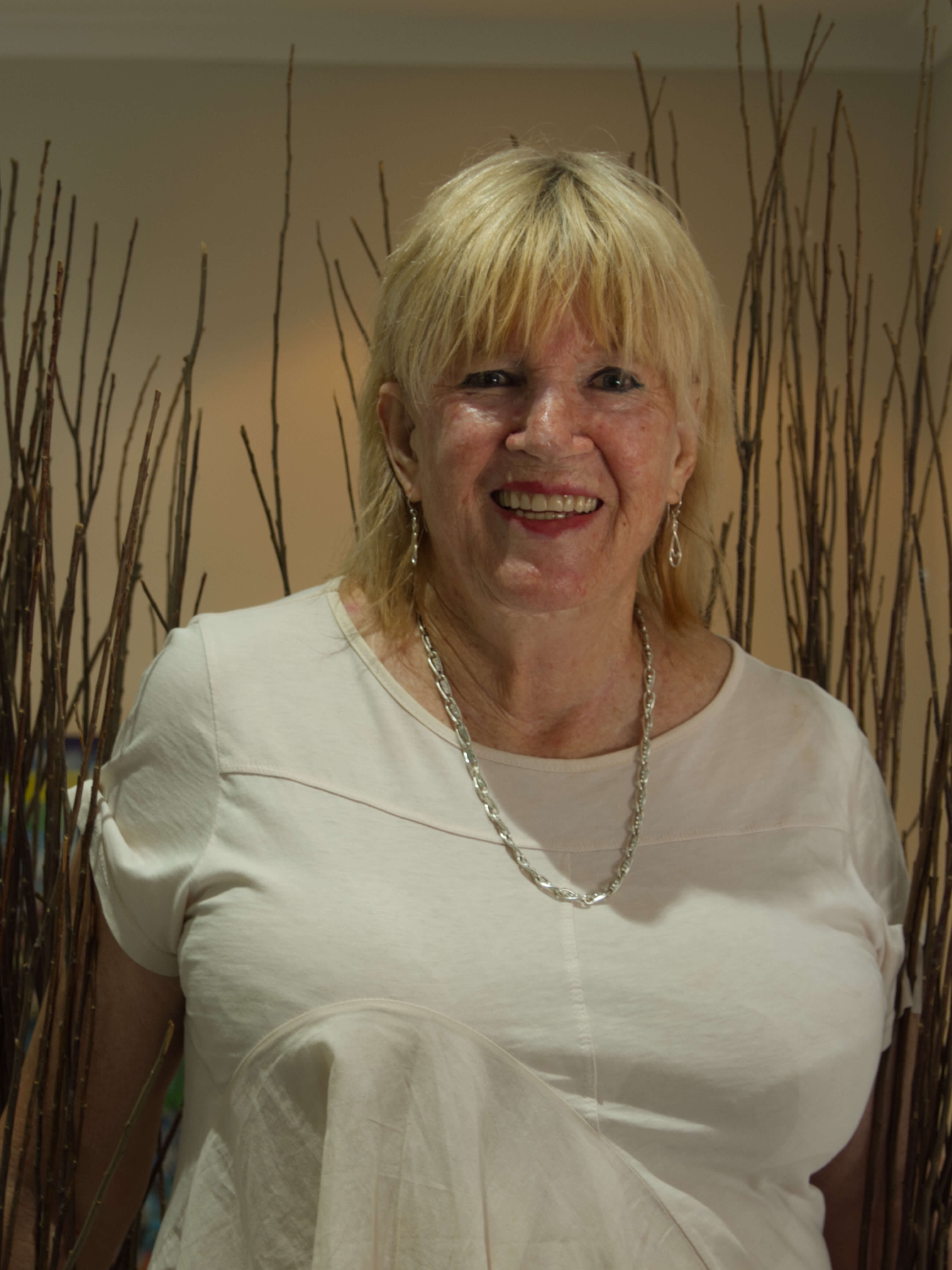 photographic portrait of a woman with shoulder length blonde hair with a fringe, gold earrings and necklace, red lipstick, white short sleeved shirt, with a brown background