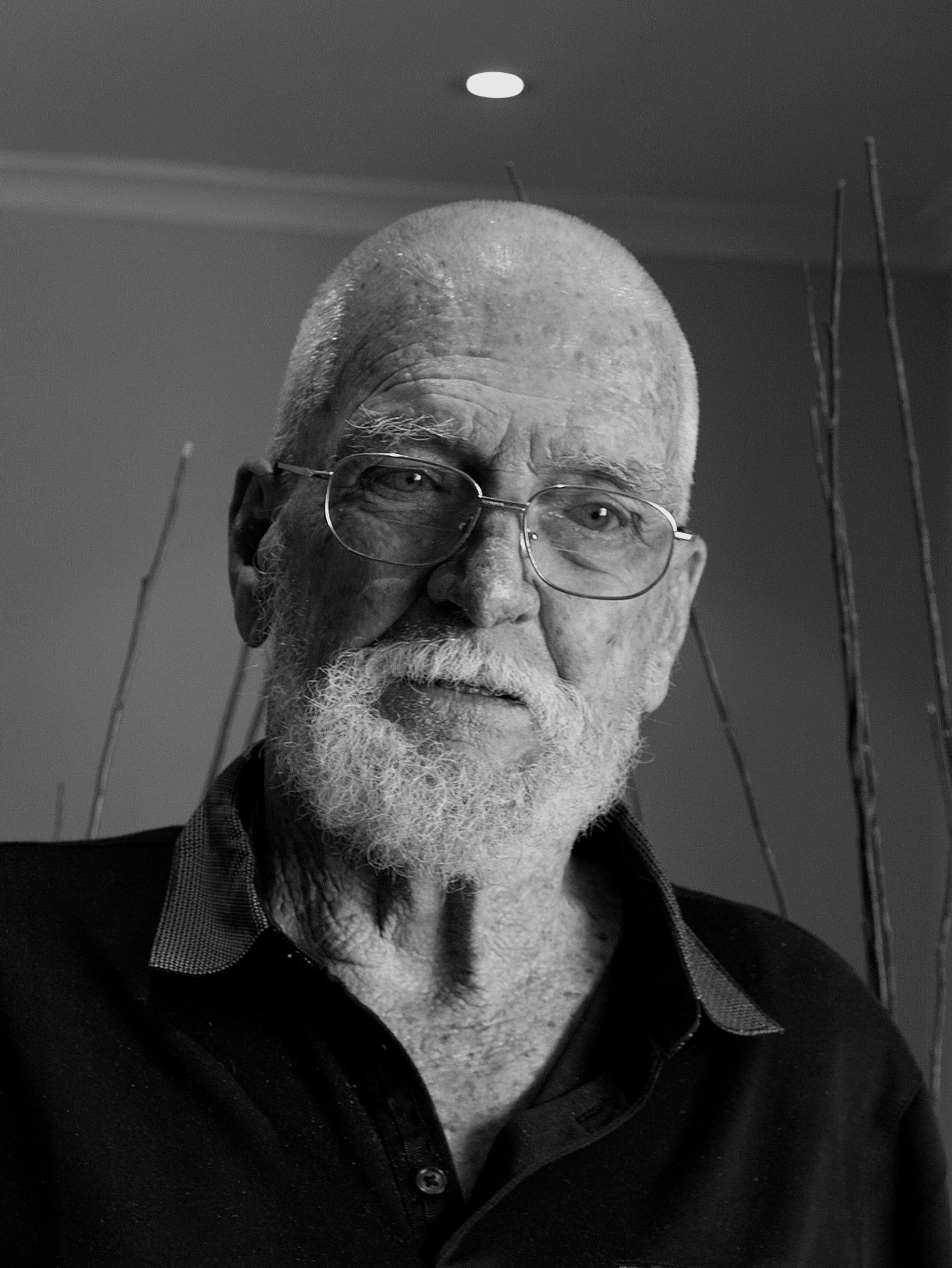 black and white photographic portrait of an elderly man, with white hair and beard, glasses and a black polo shirt.