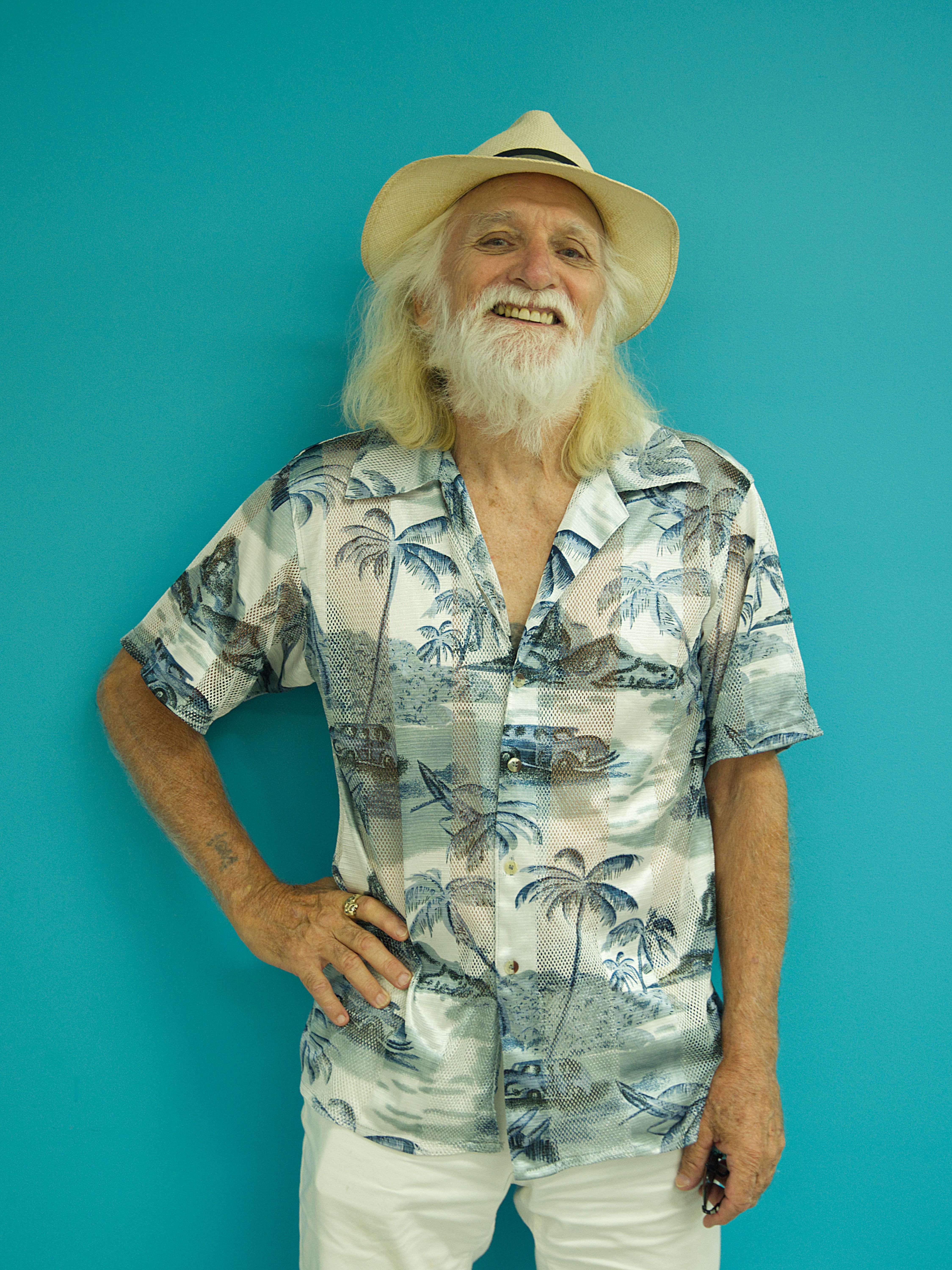 photographic portrait of a man standing against a blue background, wearing a straw hat, green and white shirt with palm trees, and white shorts. He has long white hair and a beard. He is smiling and has one had on his hip.