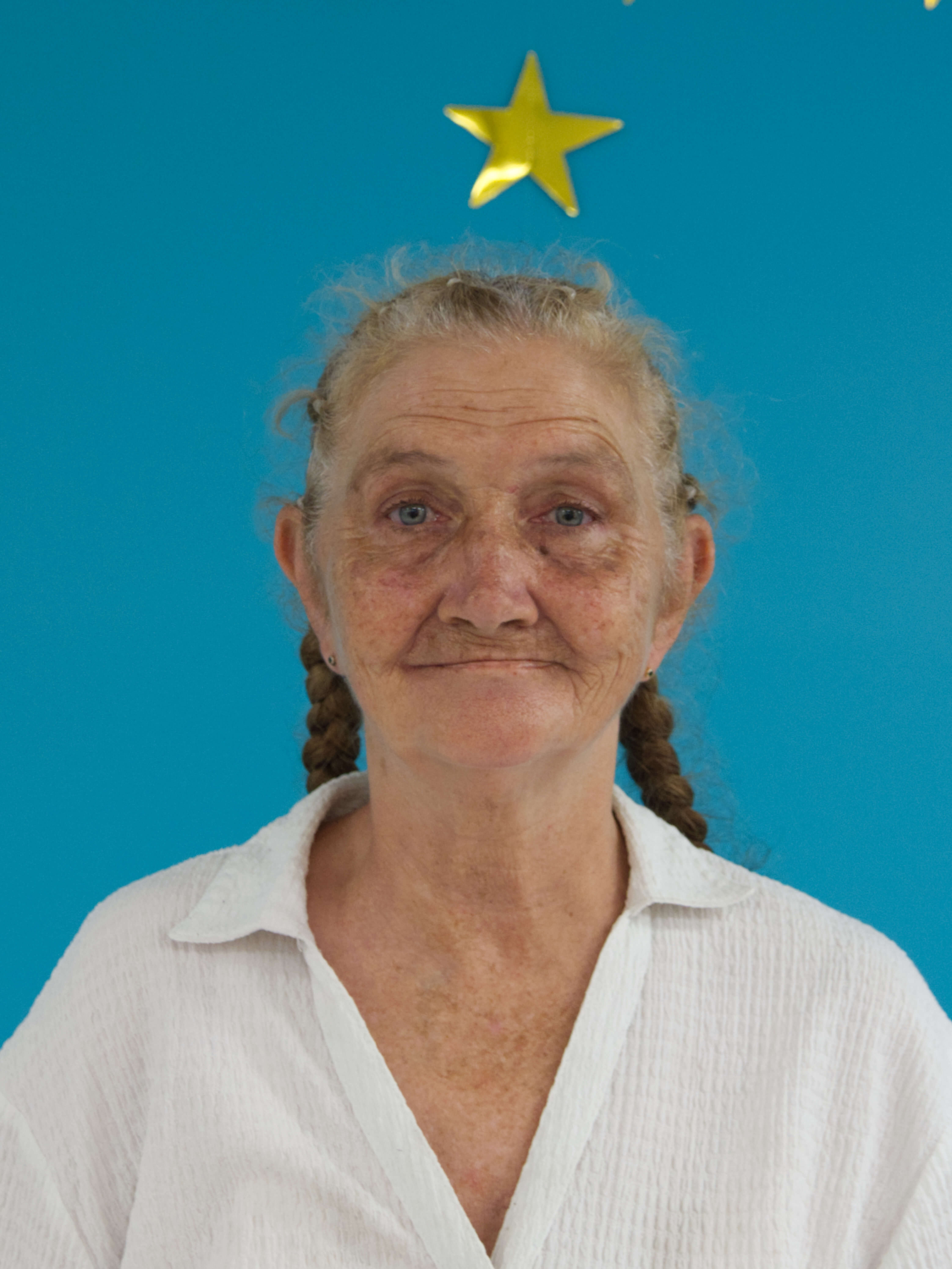 photographic portrait of a woman with a blue background. A gold star on the wall behind sits over her head. She has brown and grey hair plaited into pigtails, blue eyes and a white collared shirt