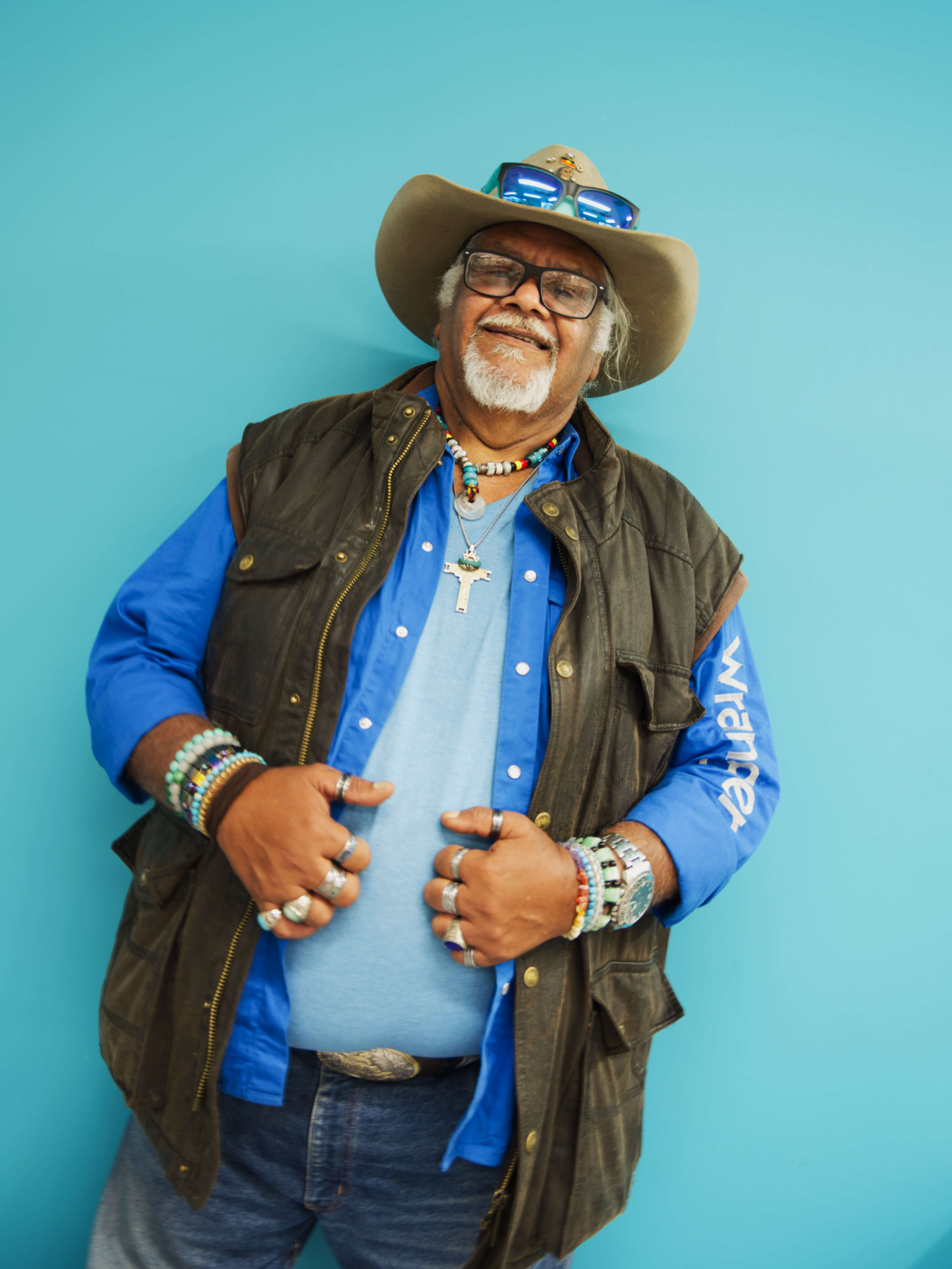 A photographic portrait of a man wearing a blue shirt, brown jacket, a cowboy hat with sun glasses on top, and jeans. He is wearing glasses, necklaces, rings and bracelets, he has white hair and a beard