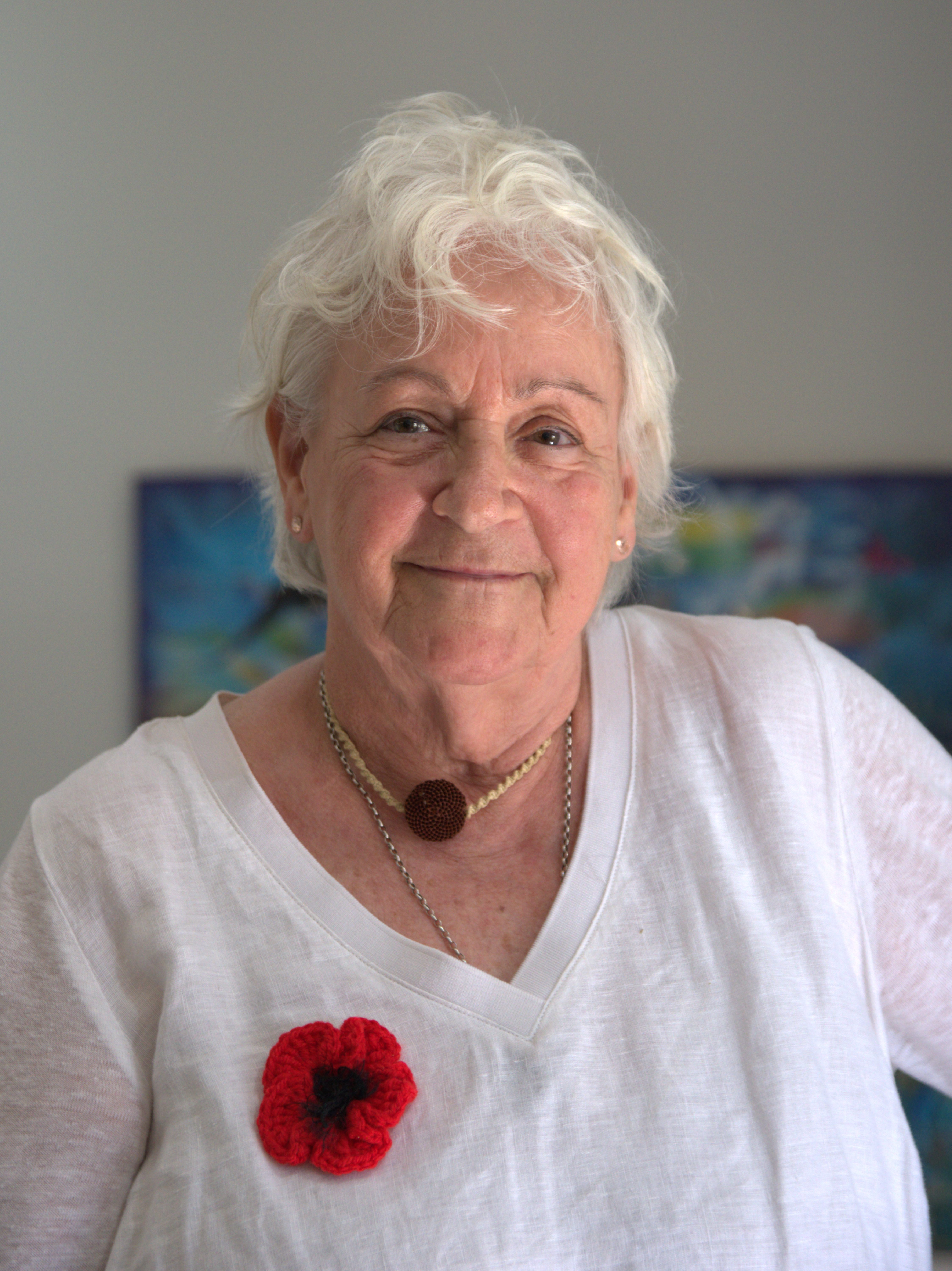 photographic portrait of a woman with white hair, green eyes, smiling at the camera, she is wearing a white t-shirt with a red poppy broach, and necklaces and earrings