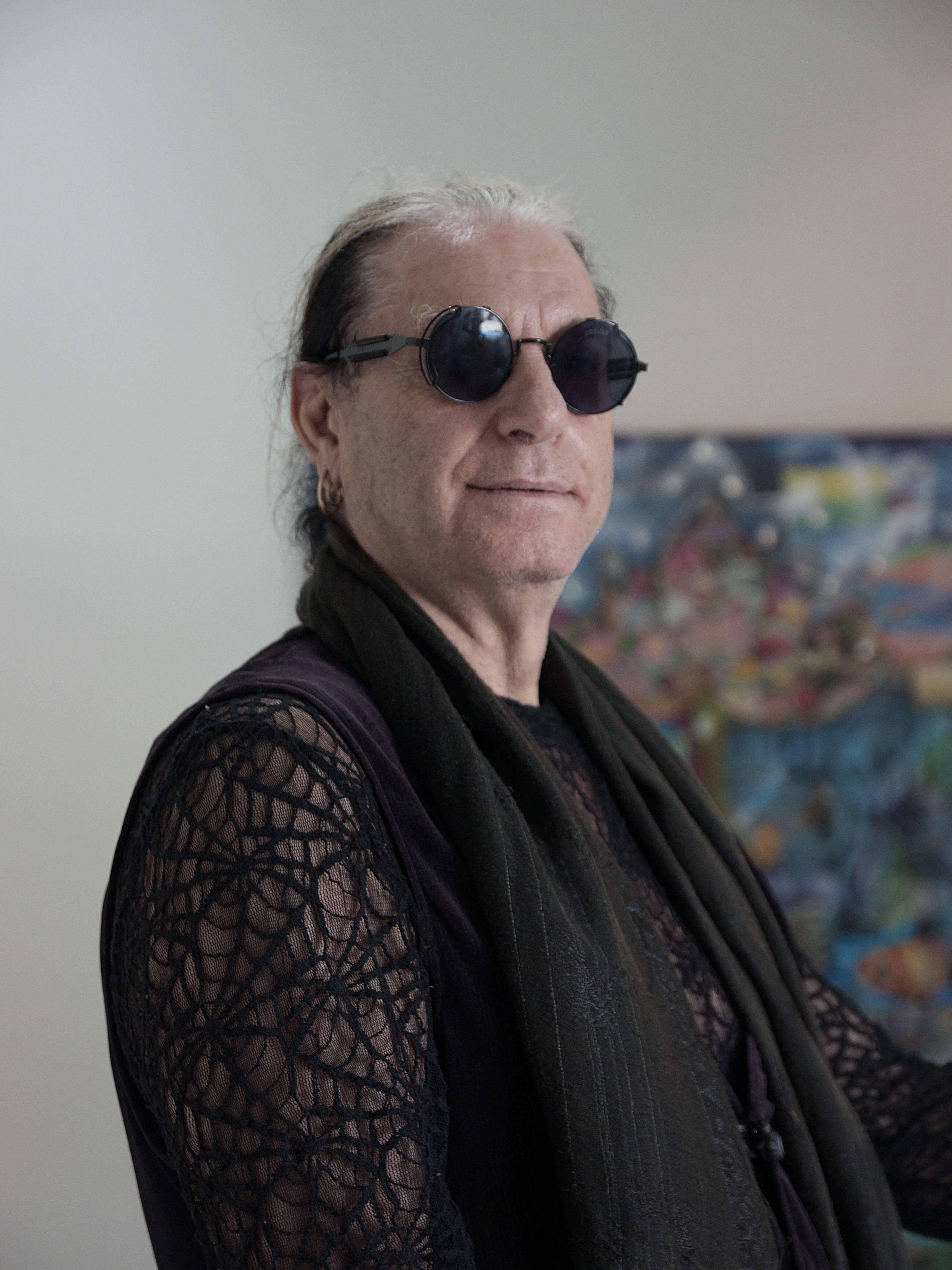 portrait of a man with white and black hair in a pony tail, he is wearing all black clothes with mesh sleeves, round sunglasses, and a gold earring
