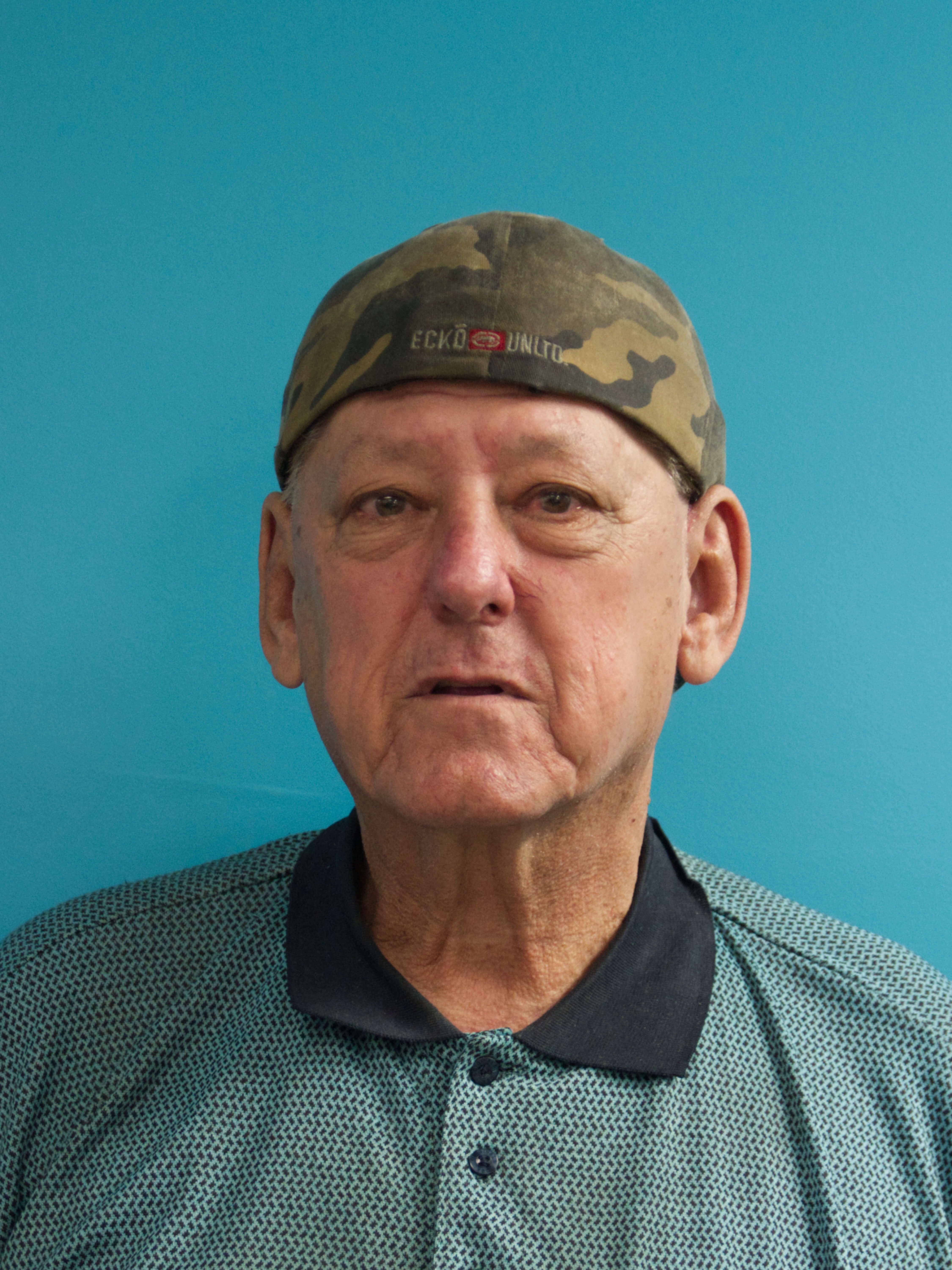 photographic portrait of a middle aged man looking at the camera, he is in front of a blue background, wearing a blue collared shirt and a backwards camo cap hat