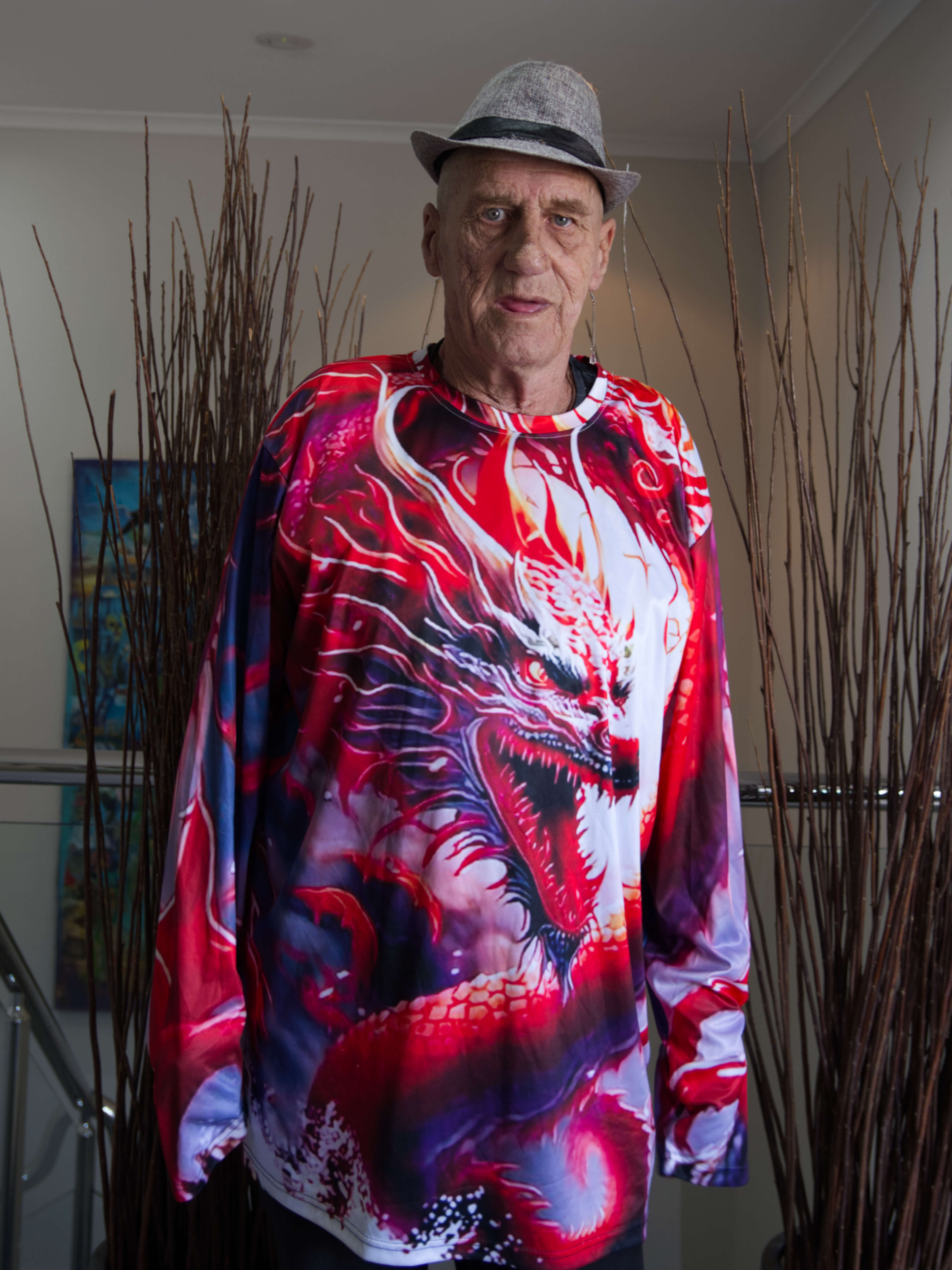 photographic portrait of a man wearing a brightly coloured shirt with a red an purple dragon. he is wearing a grey hat and behind him is a white wall and plants