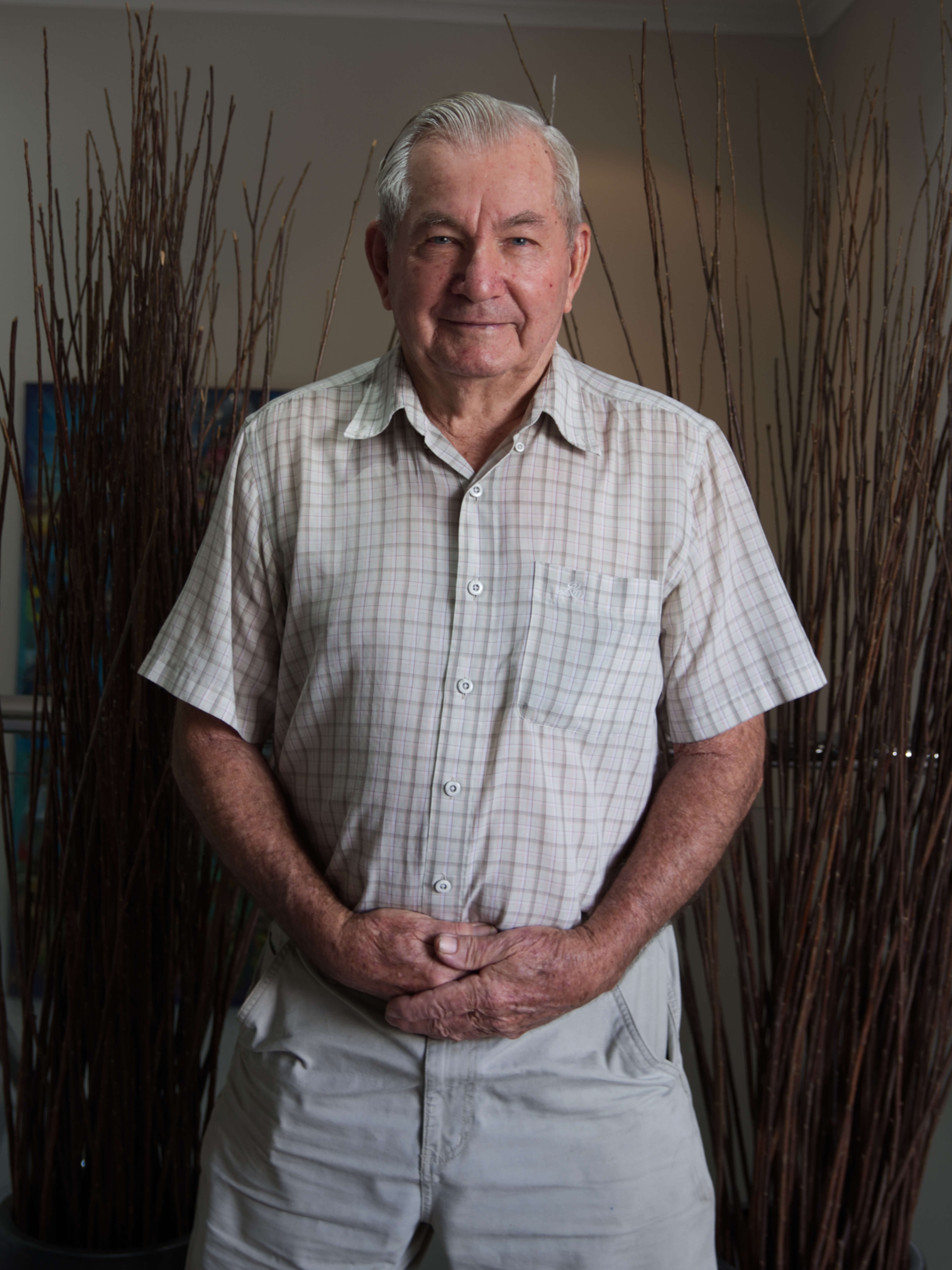photographic portrait of a older man, smiling at the camera with hands clasped at the front, wearing a white plaid shirt and white pants