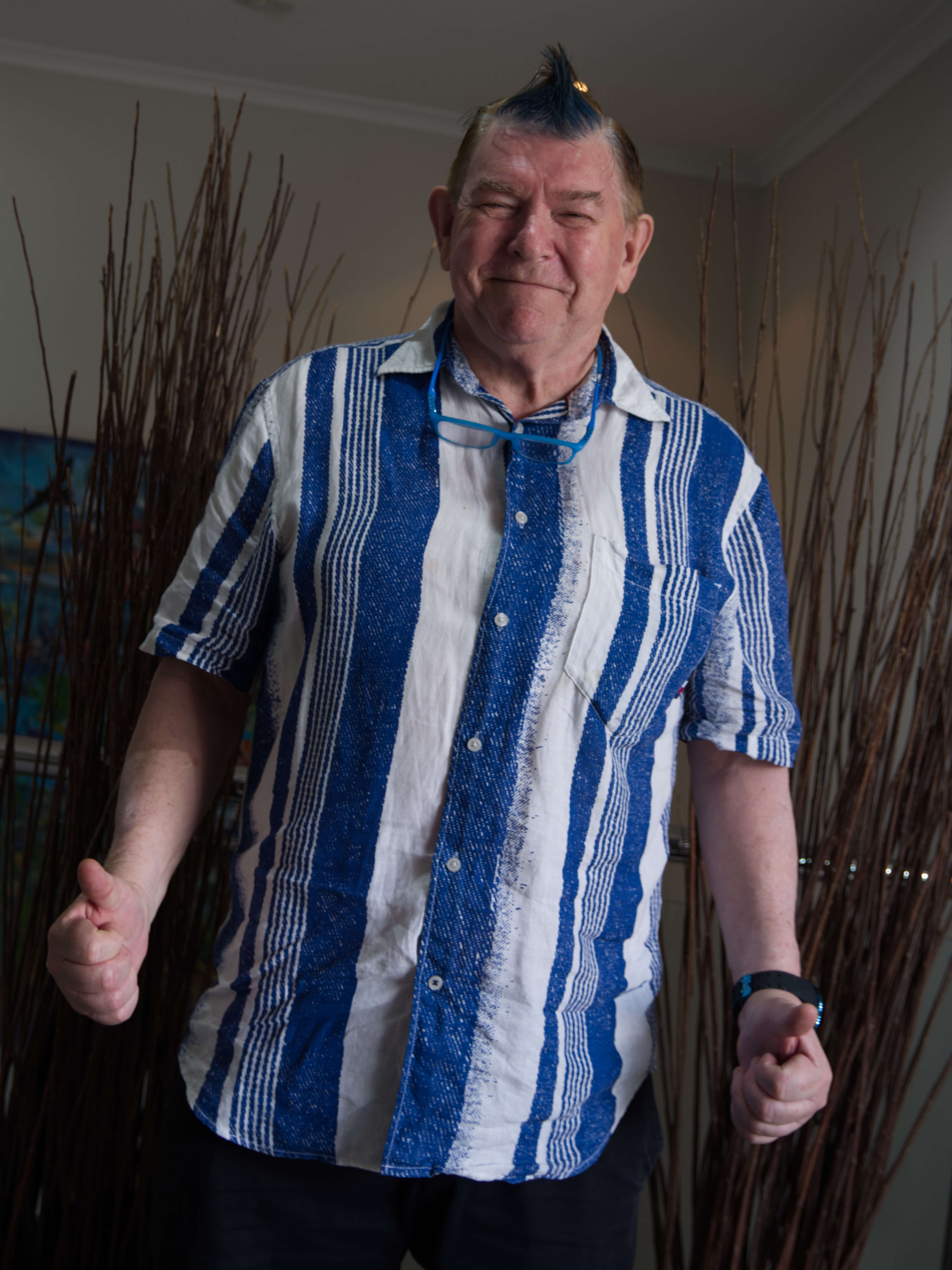 photographic portrait of a man smiling at the camera and giving the thumbs up with two hands, he has brown hair in mowhawk style and wearing a blue and white striped shirt