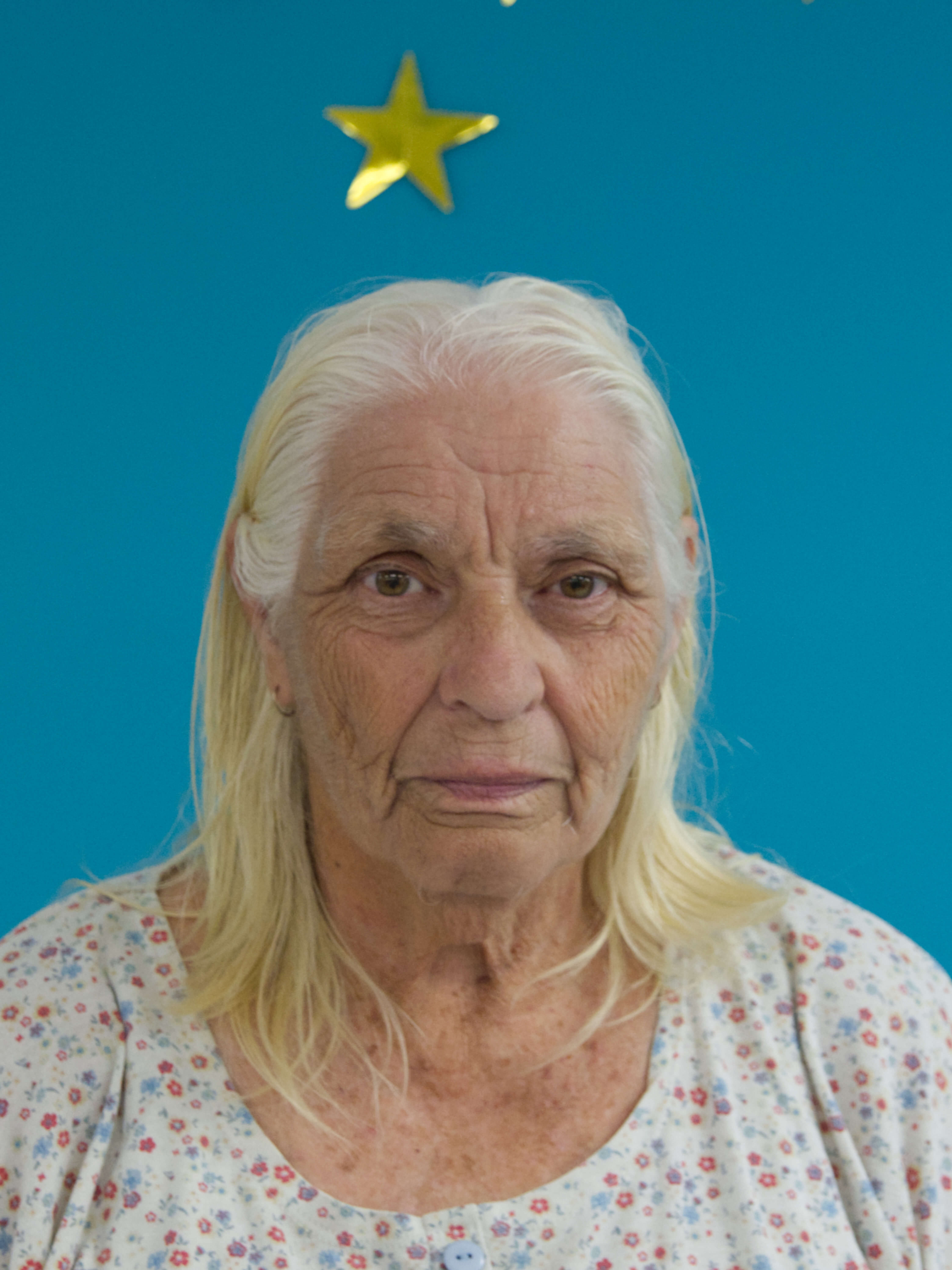 photographic portrait of an older woman with grey yellow hair, looking directly at the camera, wearing a shirt with small coloured flowers against a blue background with a gold star above her head