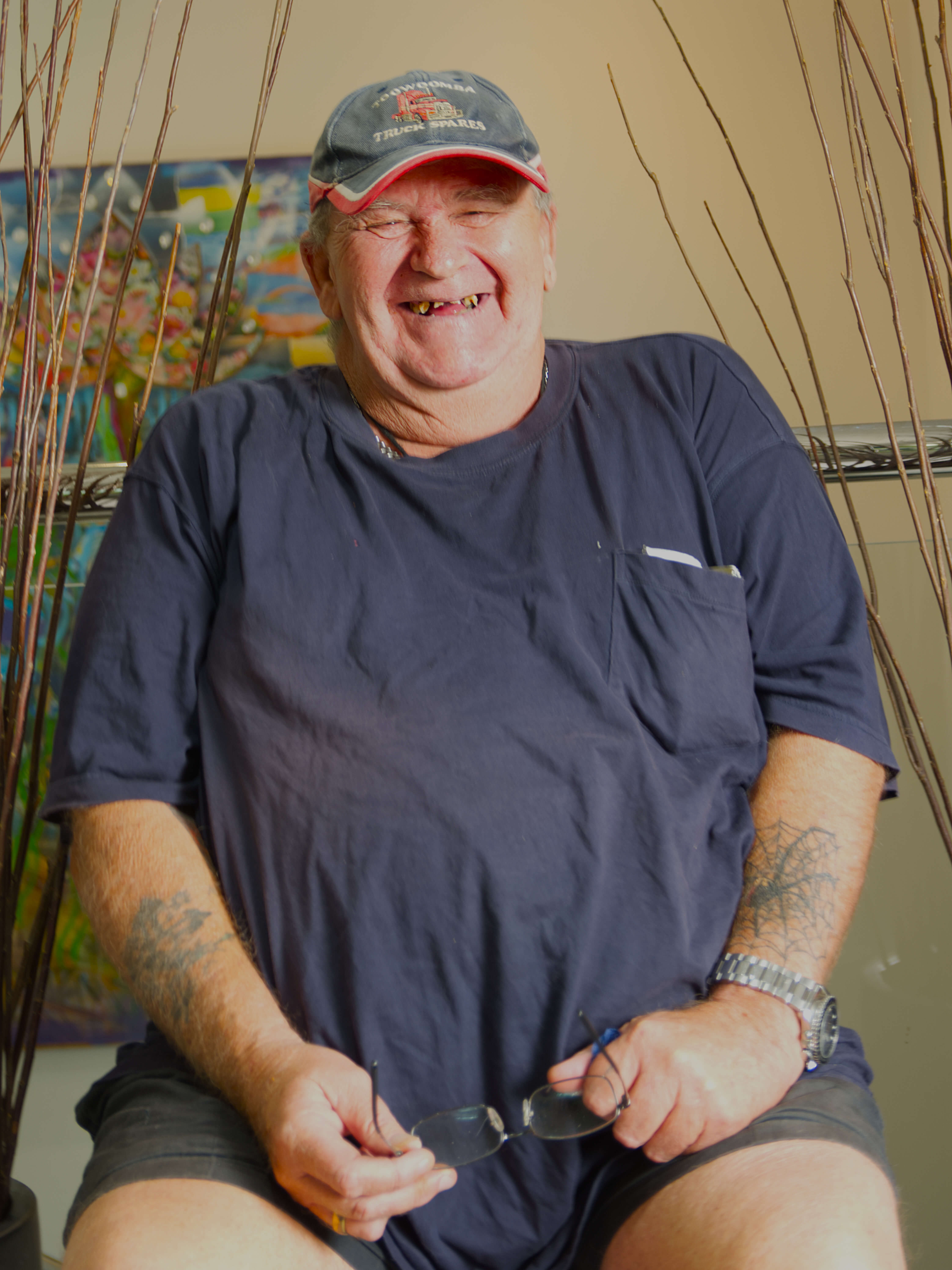 a photographic portrait of Robert Keasey, a man wearing a hat and blue shirt sitting of a chair, he has faded tattoos and a silver watch