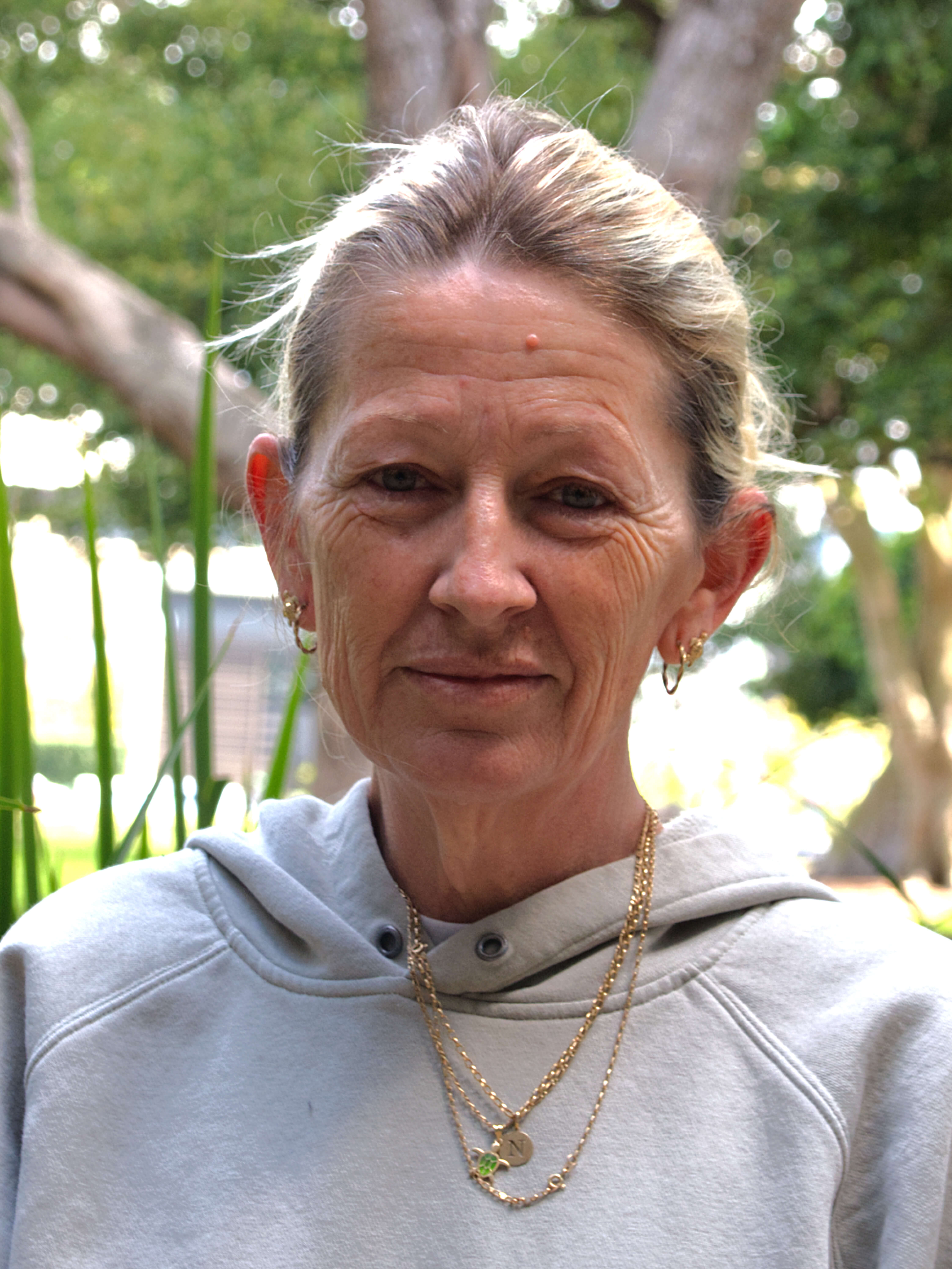 photographic portrait of Noelene Kriwat, a middle aged woman with blond hair, gold earrings and gold necklaces wearing a grey hooded jumper