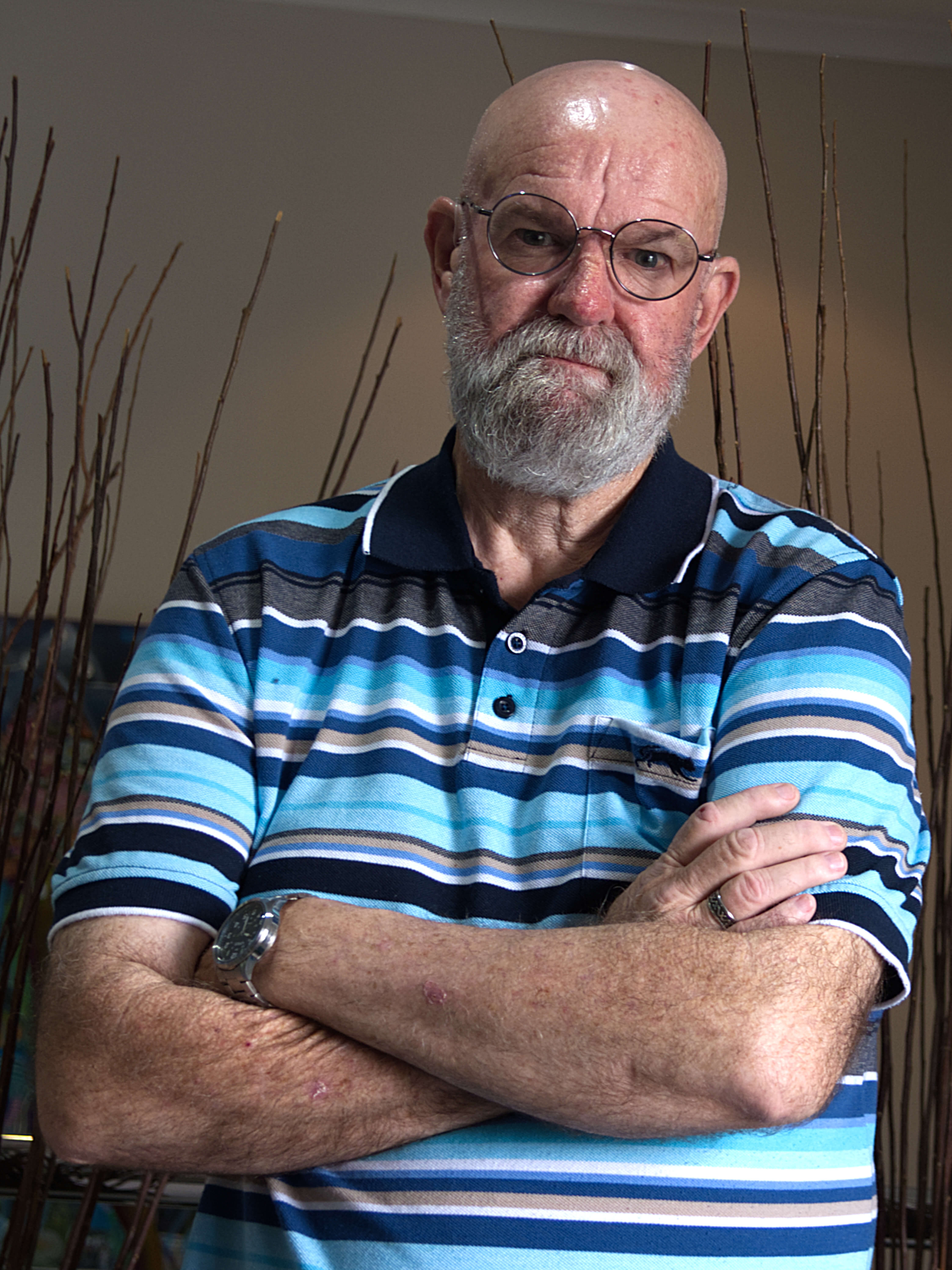 photographic portrait of Colin Lofgren, a old man with bald head, glasses and grey beard, looking sternly at the camera, wearing striped blue and white polo shirt