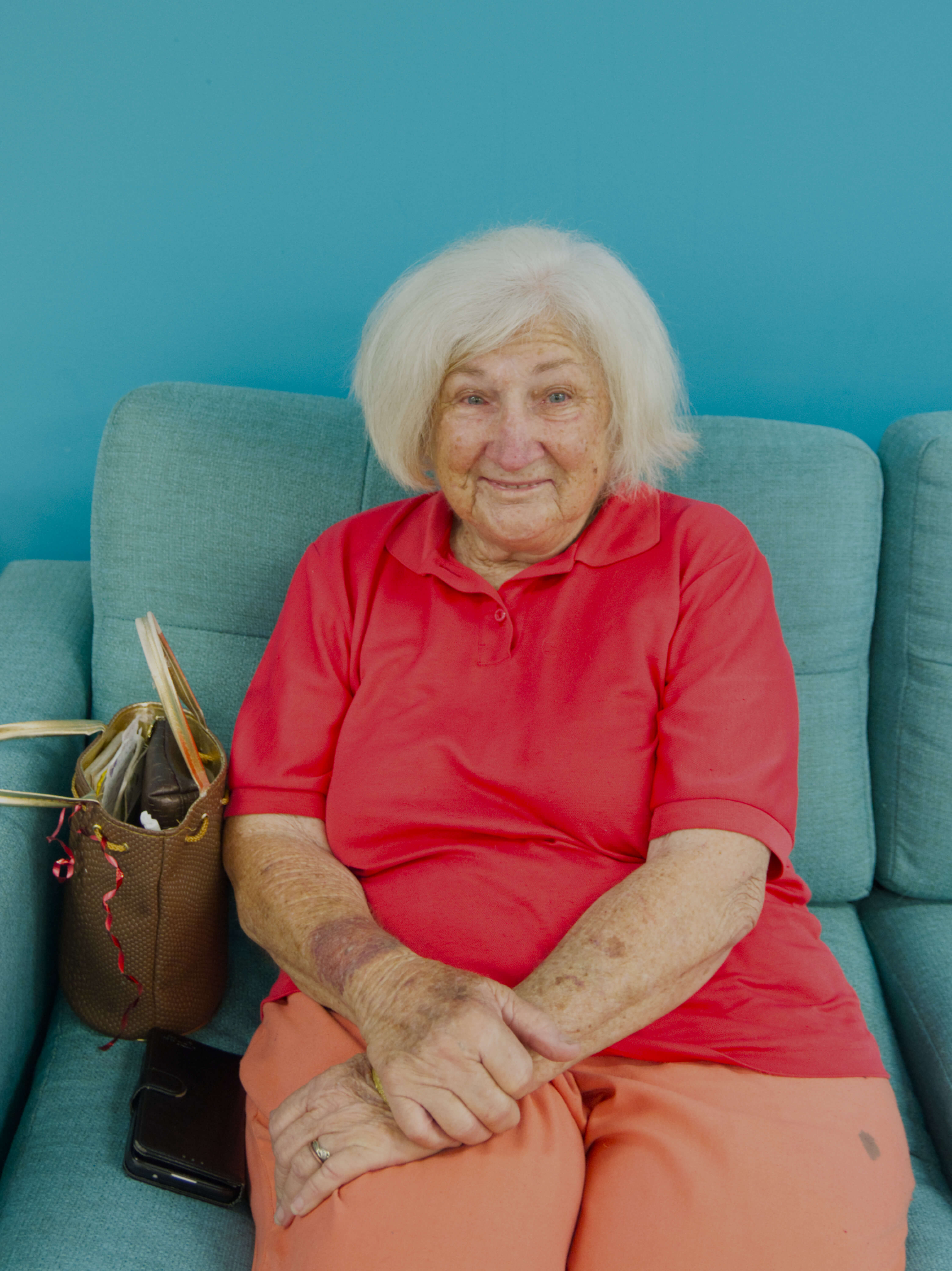 photographic portrait of Gloria Lovely, older woman with shoulder length white hair, wearing a red polo shirt, and orange pants sitting on a blue couch, next to her is a brown handbag