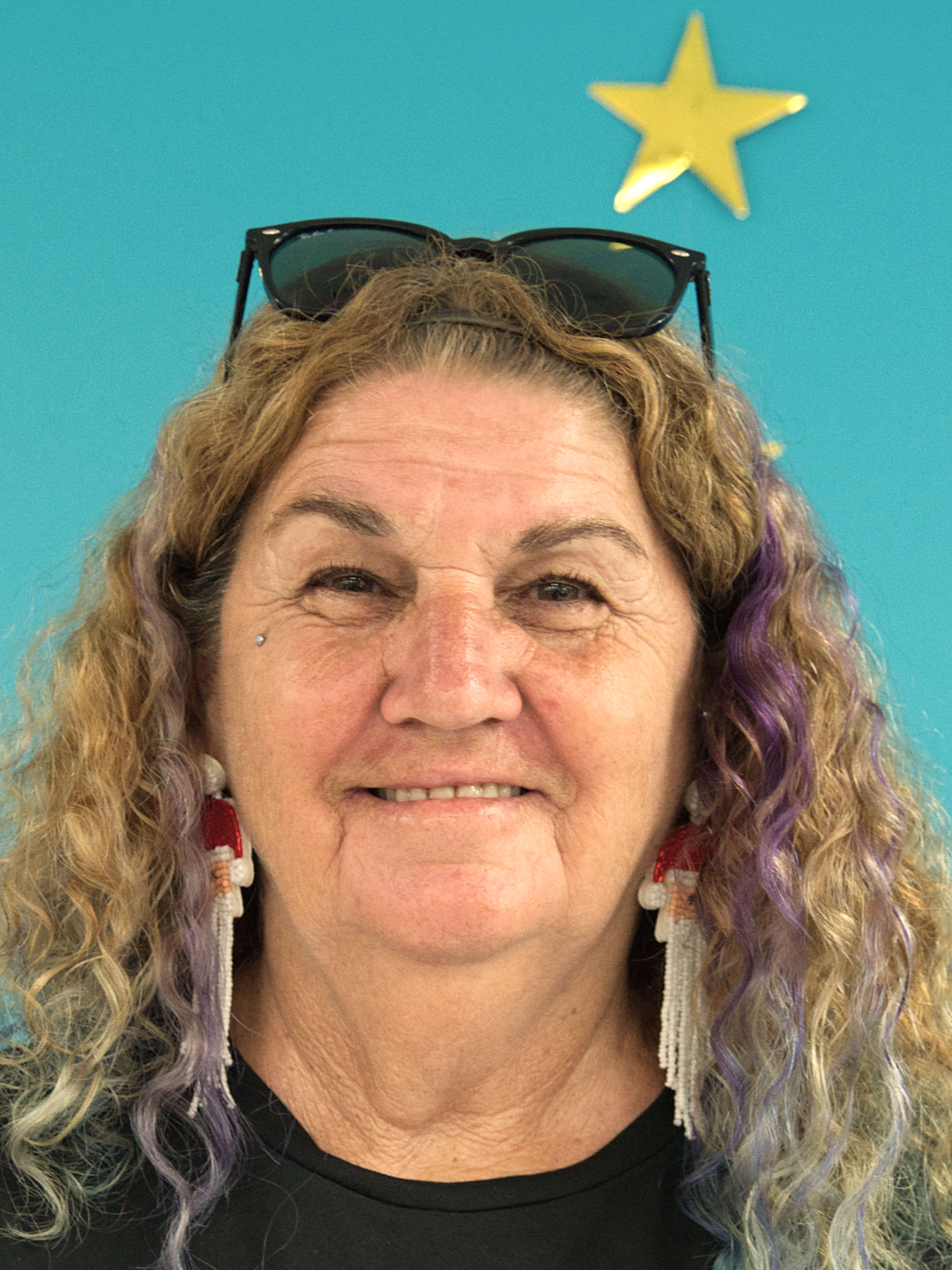 photographic portrait of Karen Riding, she has blonde and purple hair, sunglasses, earrings and a gold star above her head, she is smiling at the camera on a blue background