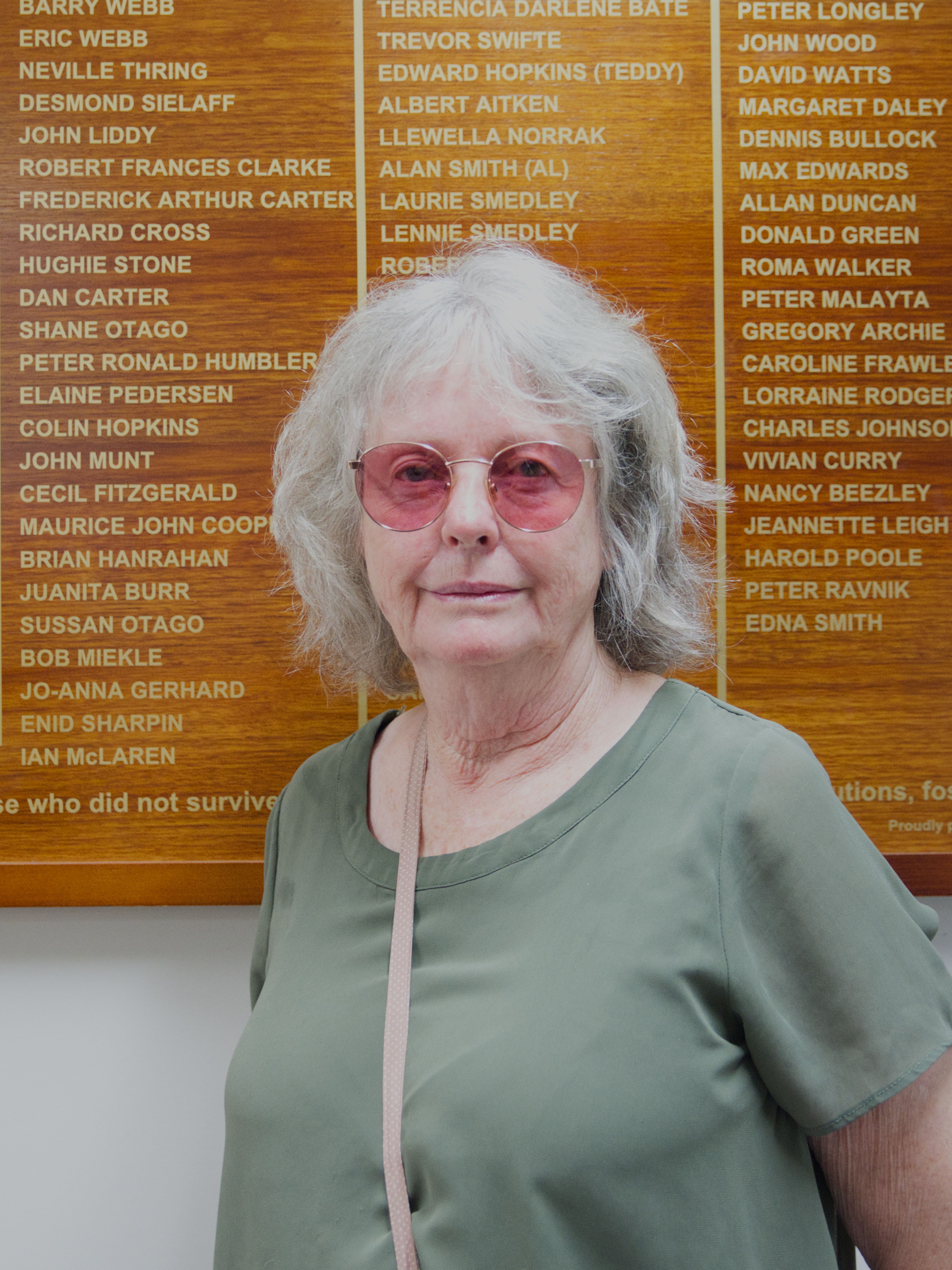 photographic portrait of Michele Urwin, older woman with pink coloured glasses, and shoulder length grey hair, wearing a green t-shirt with a bag strap across her body
