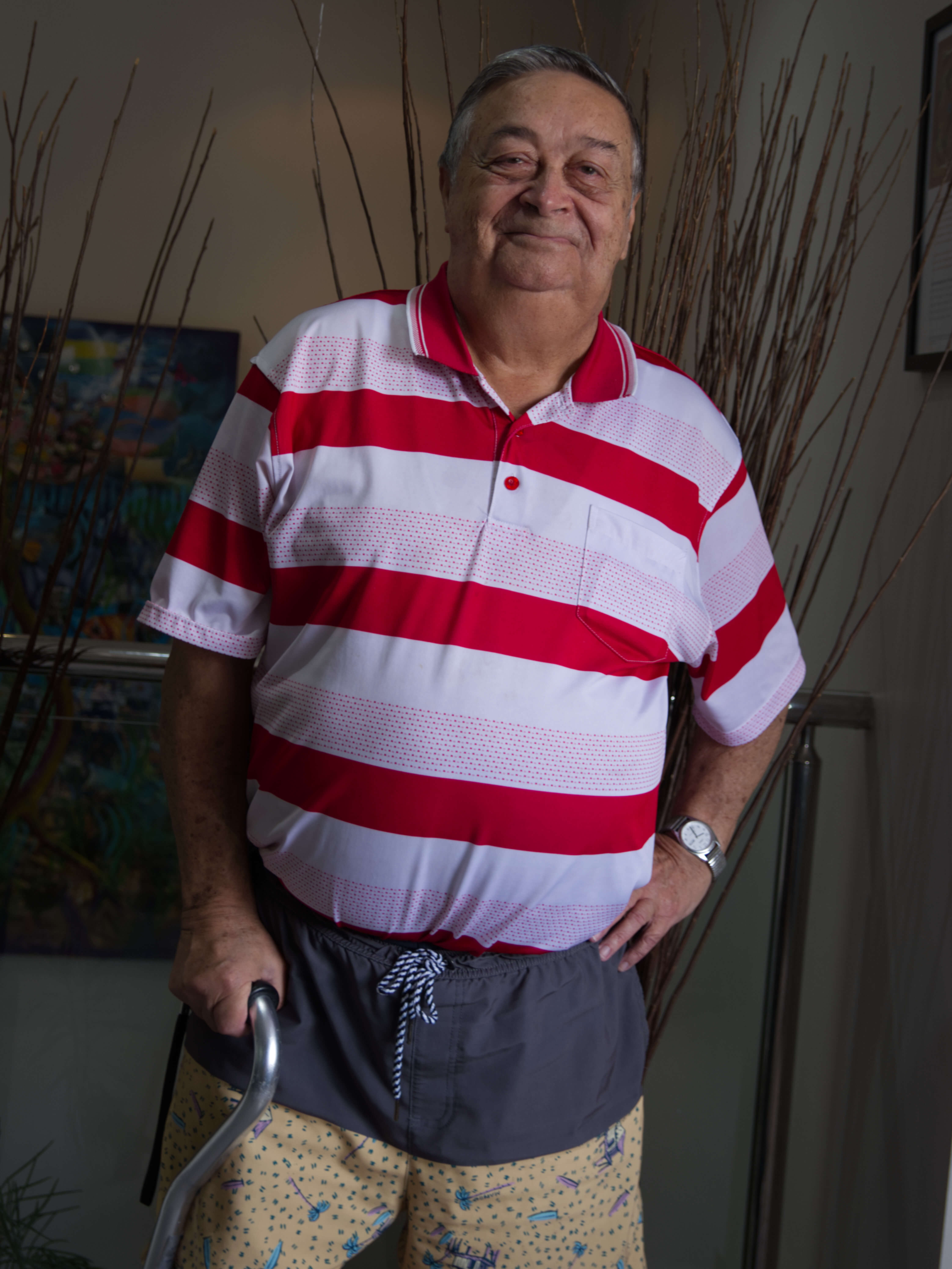 photographic portrait of Ken Waldron, he is a older man with a walking stick, wearing a red and pink striped shirt and shots, smiling at the camera