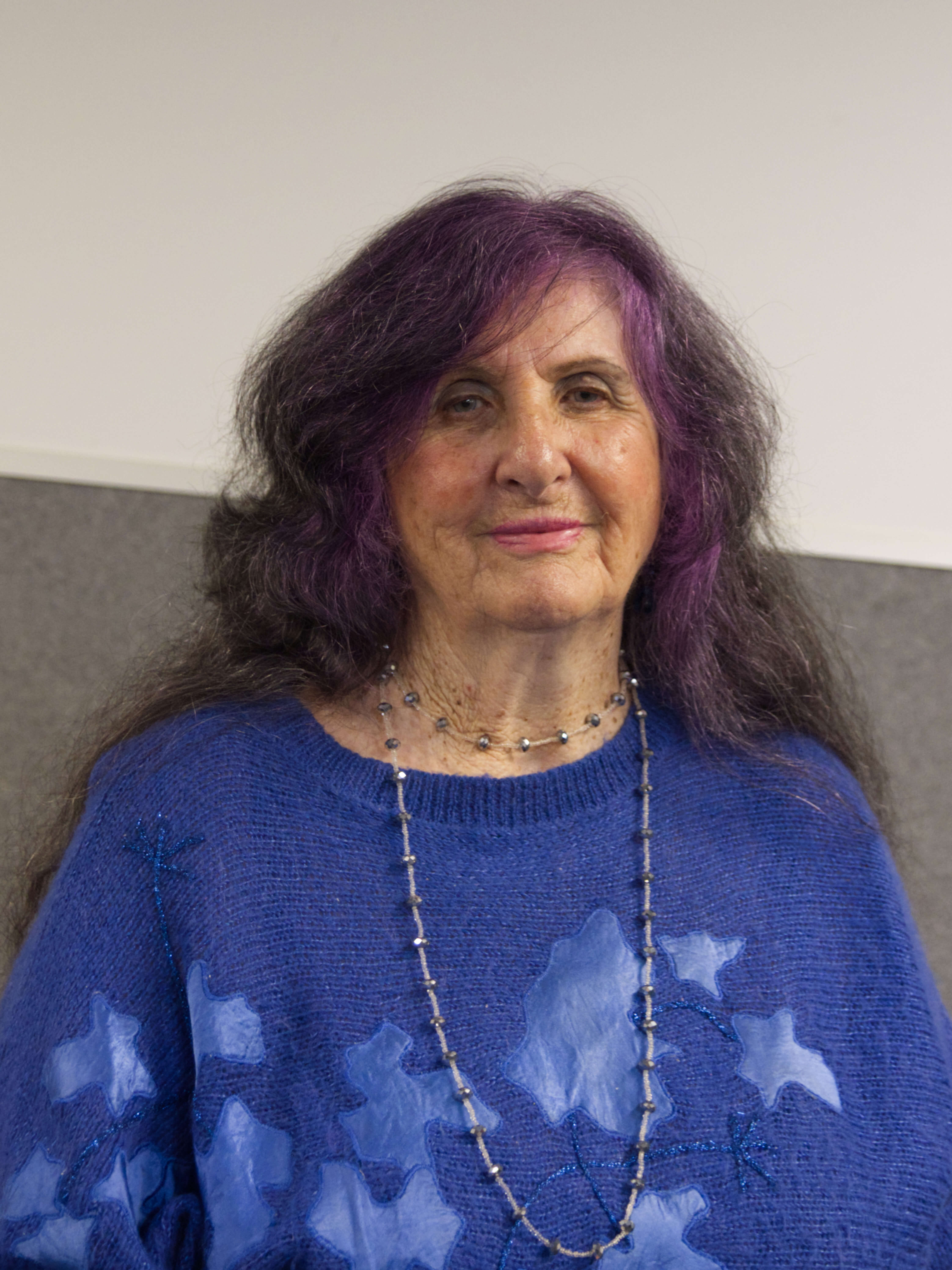 photographic portrait of Theresa Whitfield, a woman with long black and pink hair, wearing a long necklace and blue long sleeved shirt smiling at the camera