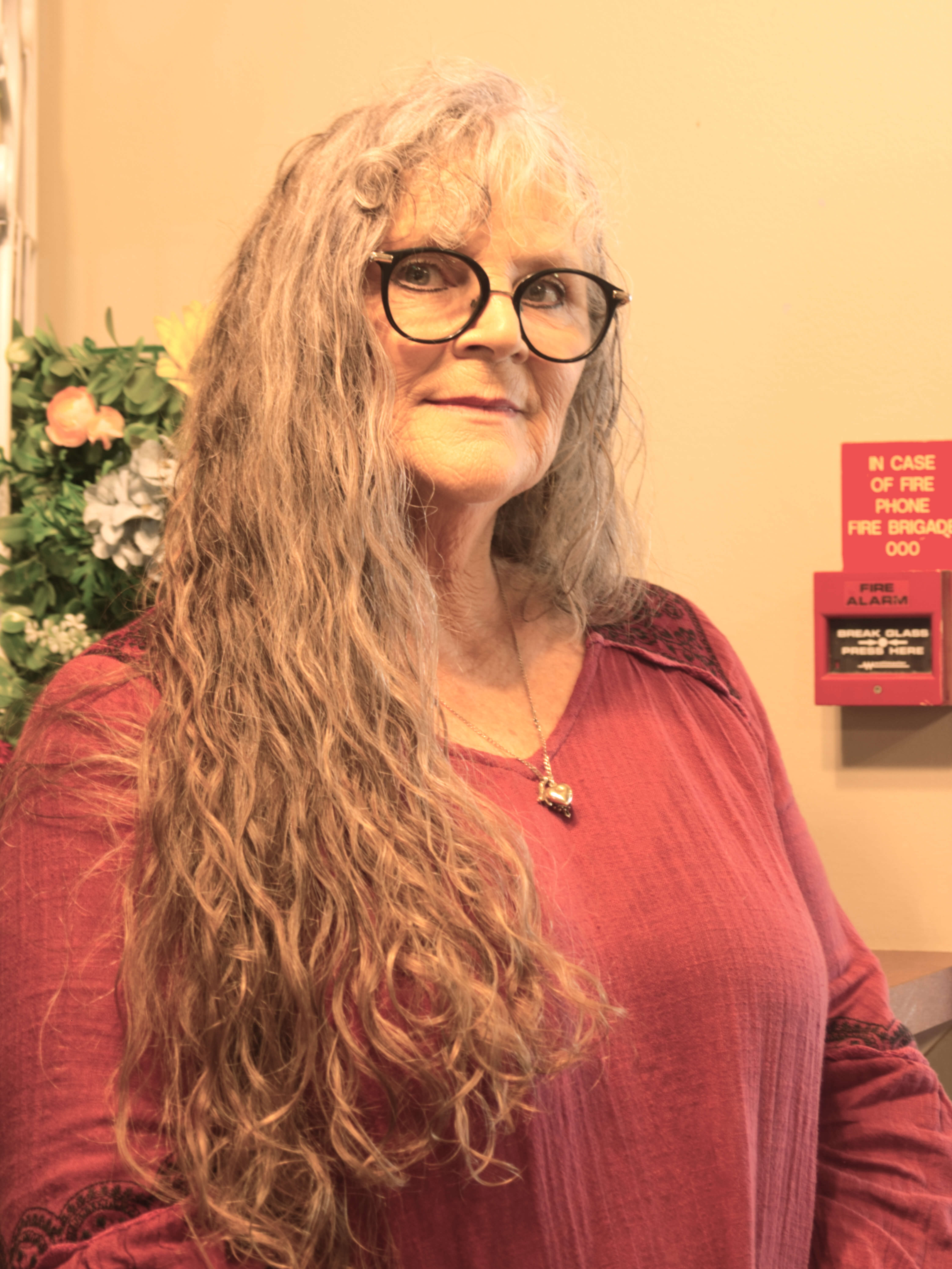 photographic portrait of Kim Wilson, a woman with long grey hair and black rimmed glasses, wearing a pink shirt and a necklace