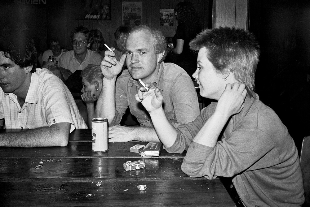 Concert goers sit at a bar with beer and cigarettes in hand.