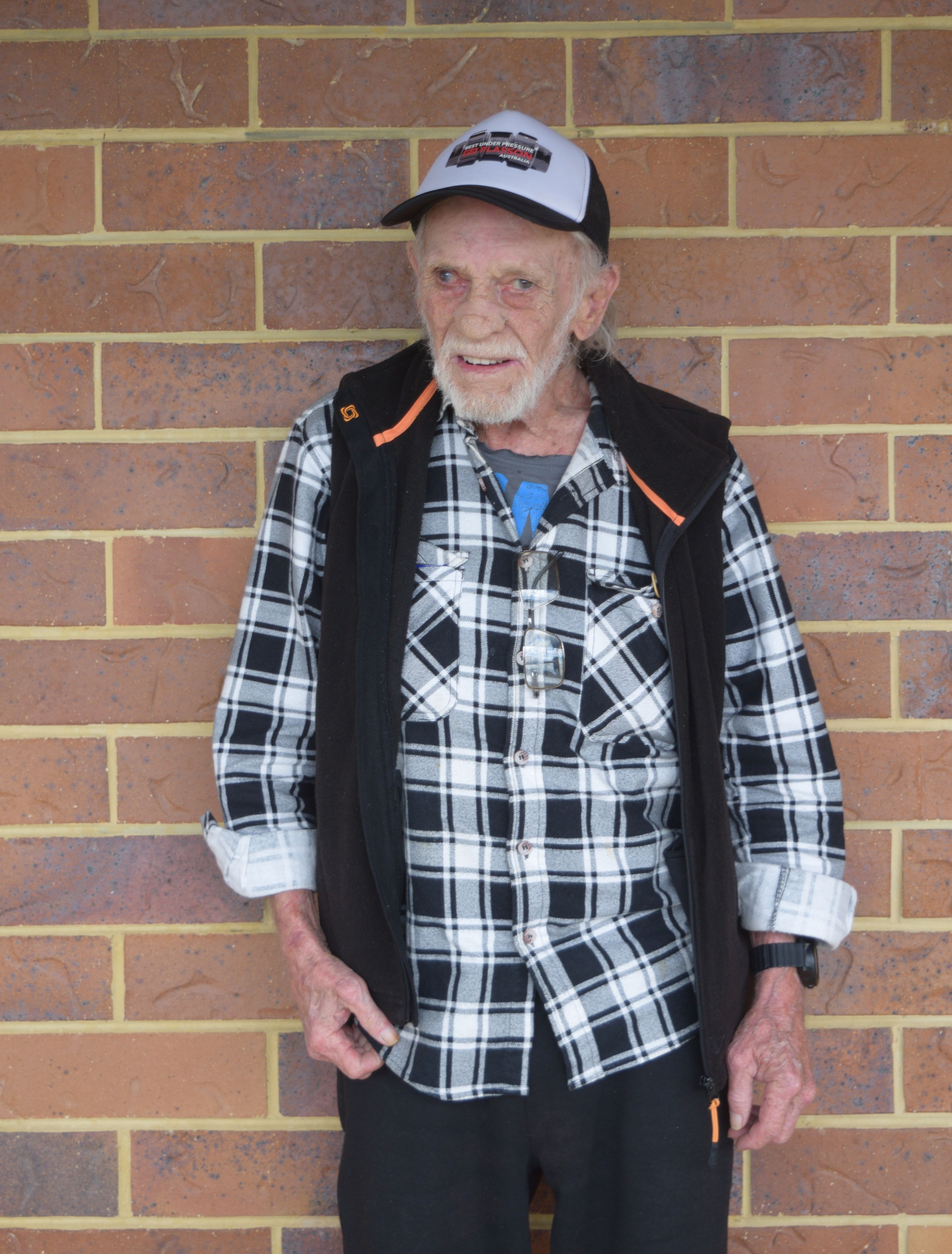 photographic portrait of Harold James, older man with great hair standing against a brick wall, wearing black and white plaid shirt and black vest 