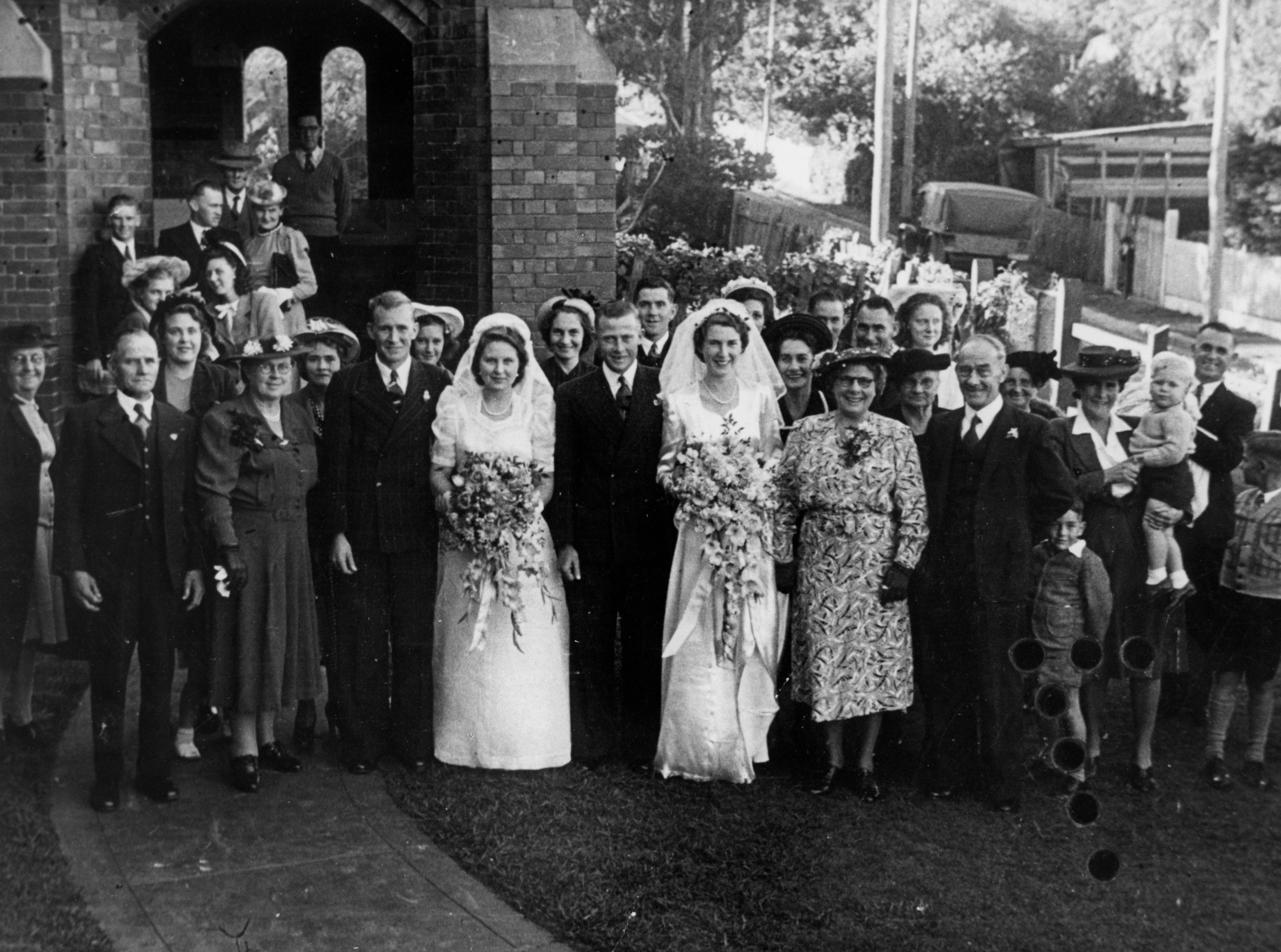 Double wedding in East Brisbane 1949 Two brides with large bouquets stand next to their grooms in front of a large group of people oustide a church