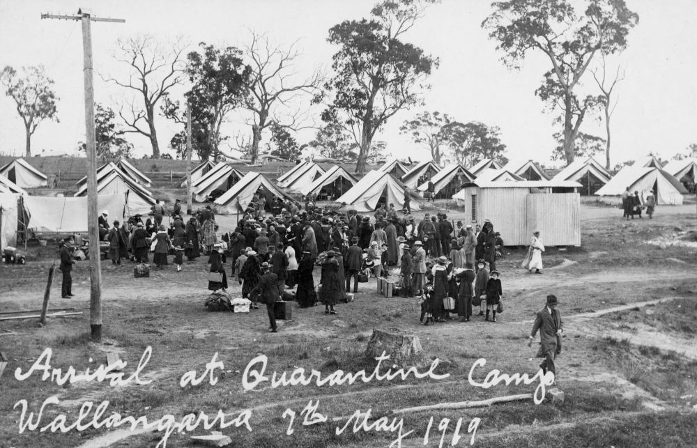 People arrive at the quarantine camp in Wallangarra during the influenza epidemic of 1919. The camp consists of tents and corrugated iron buildings, surrounded by bushland. People are waiting to be registered and admitted to the camp. They have their suitcases and other belongings with them.