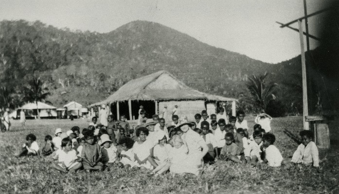 A group of children sat on the ground in front of a building with a roof made of dried straw 