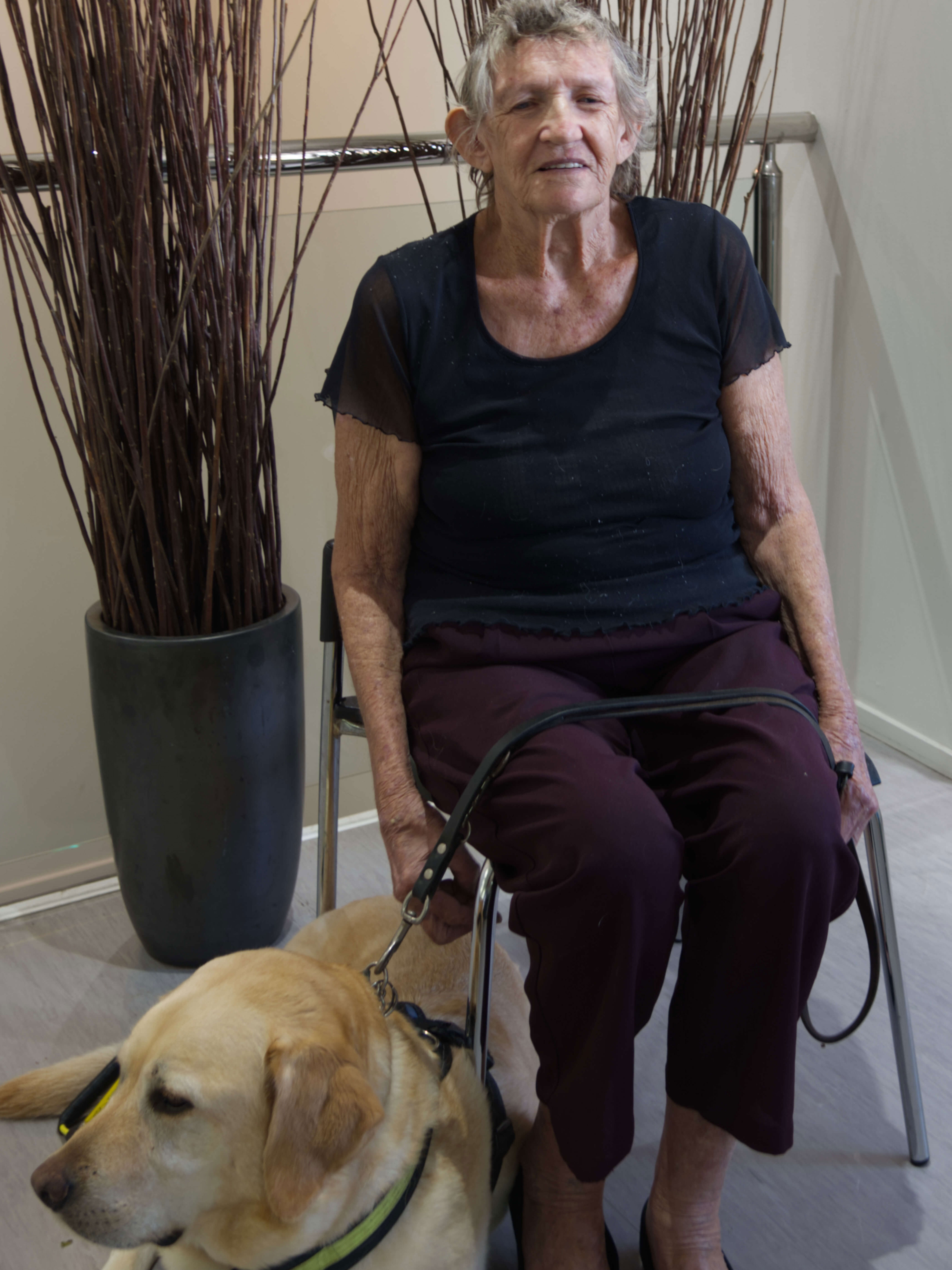 Portrait of Margaret Hourigan smiling at the camera. She is wearing black clothes and sitting on a chair, she is holding the lead of her guide dog.