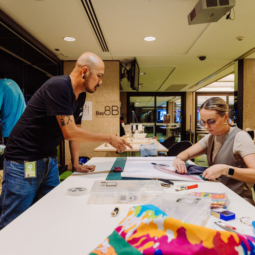 People sewing at The Edge during Threads 2024, someone is visible on the right hand side using a ruler to cut out a pattern, someone on the left hand side is standing over the table and pointing at the pattern to help.