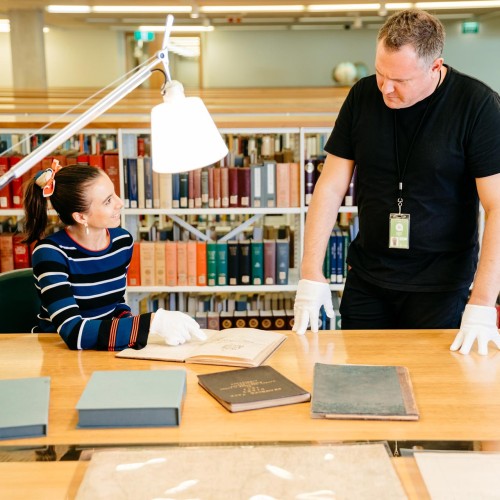 Visitor talking to State Library staff member. The visitor is wearing white gloves while pointing to old books.