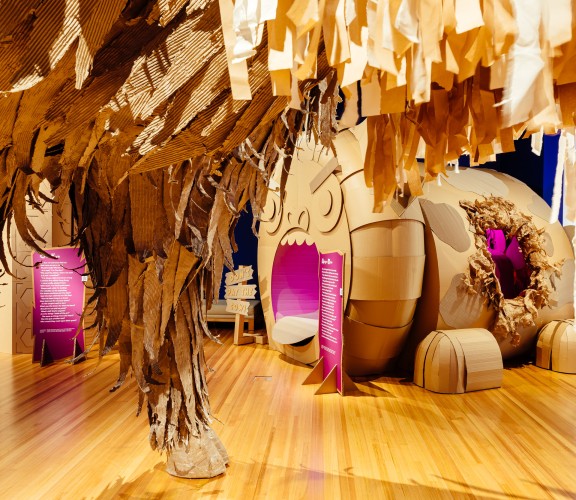A large cardboard leg from an imaginary beast in the foreground with a cardboard cow in the background who has a very animated face.