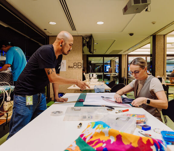 People sewing at The Edge during Threads 2024, someone is visible on the right hand side using a ruler to cut out a pattern, someone on the left hand side is standing over the table and pointing at the pattern to help.