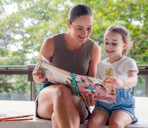 Ash Barty reading a book to a young girl outside at State Library of Queensland