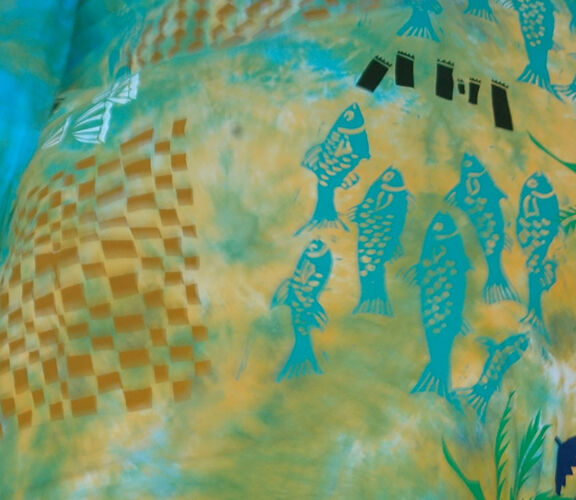 A child holding up a large, colourful fabric decorated with fish, palm trees, woven patterns, and abstract shapes in shades of blue, green, yellow, and orange.