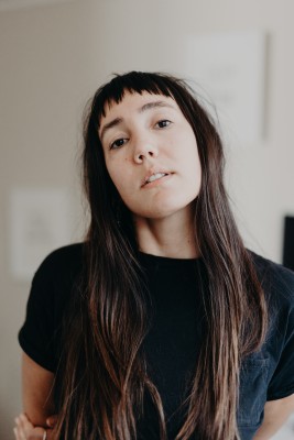 A person with long brown hair looking at the camera with head tilted back. They are wearing a black shirt against a white background.