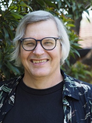 Headshot of B. R. Dionysius. He has dark rimmed glasses and stands in a garden.