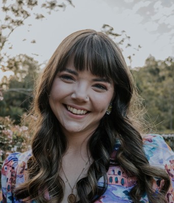 Emily Lighezzolo stands in a garden; she has on a bright shirt and dark wavy hair