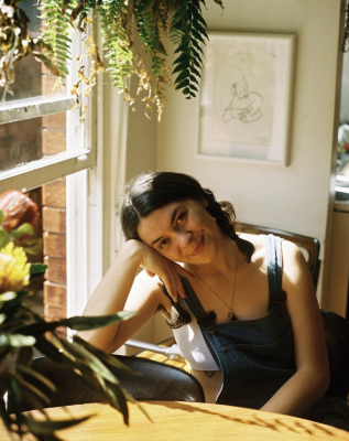 A young woman wearing overalls sits at a wooden table and smiles at the camera