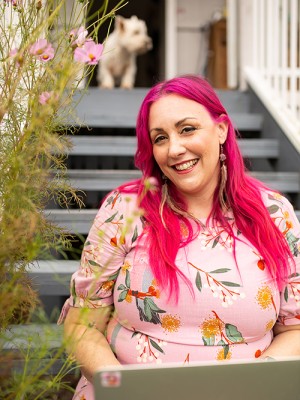 A photo of Melanie Saward sitting on the stairs of a house. She has pink hair and a bright dress and a little white dog is in the background.