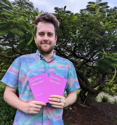 Sean West smiles and holds up two purple books. In the background are trees.