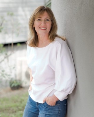 Fiona Robertson stands in a park, against a grey wall. She wears a white shirt and is smiling at the camera