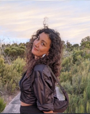 Mykaela Saunders is walking along a coastal path at sunset. She has long brown hair and big silver earrings.