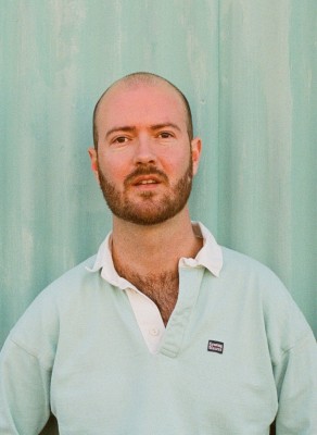 Headshot of Myles McGuire wearing a light green jumper against a green wall.