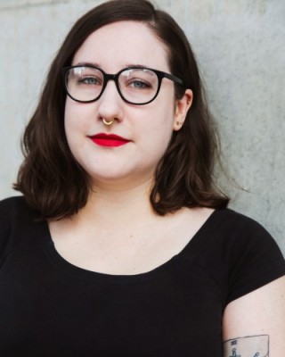 Rebecca Cheers – headshot – she wears a black t-shirt and red lipstick. Her left arm has a tattoo of a heart and swords.