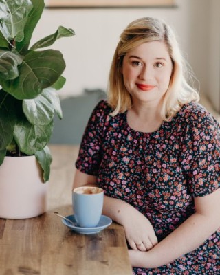 Rhiannon Wilde sits at a desk next to a fiddle leaf fig. She is wearing a floral dress and is smiling.