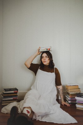 Wallea Eaglehawk sits against a white wall, with books around her, and she holds a stack on her head