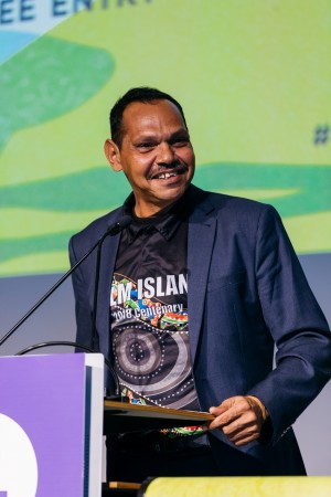 Palm Island Mayor Alf Lacey stands at a podium with a microphone. He is wearing a suit jacket and a polo shirt celebrating the 2018 Centenary of Palm Island.